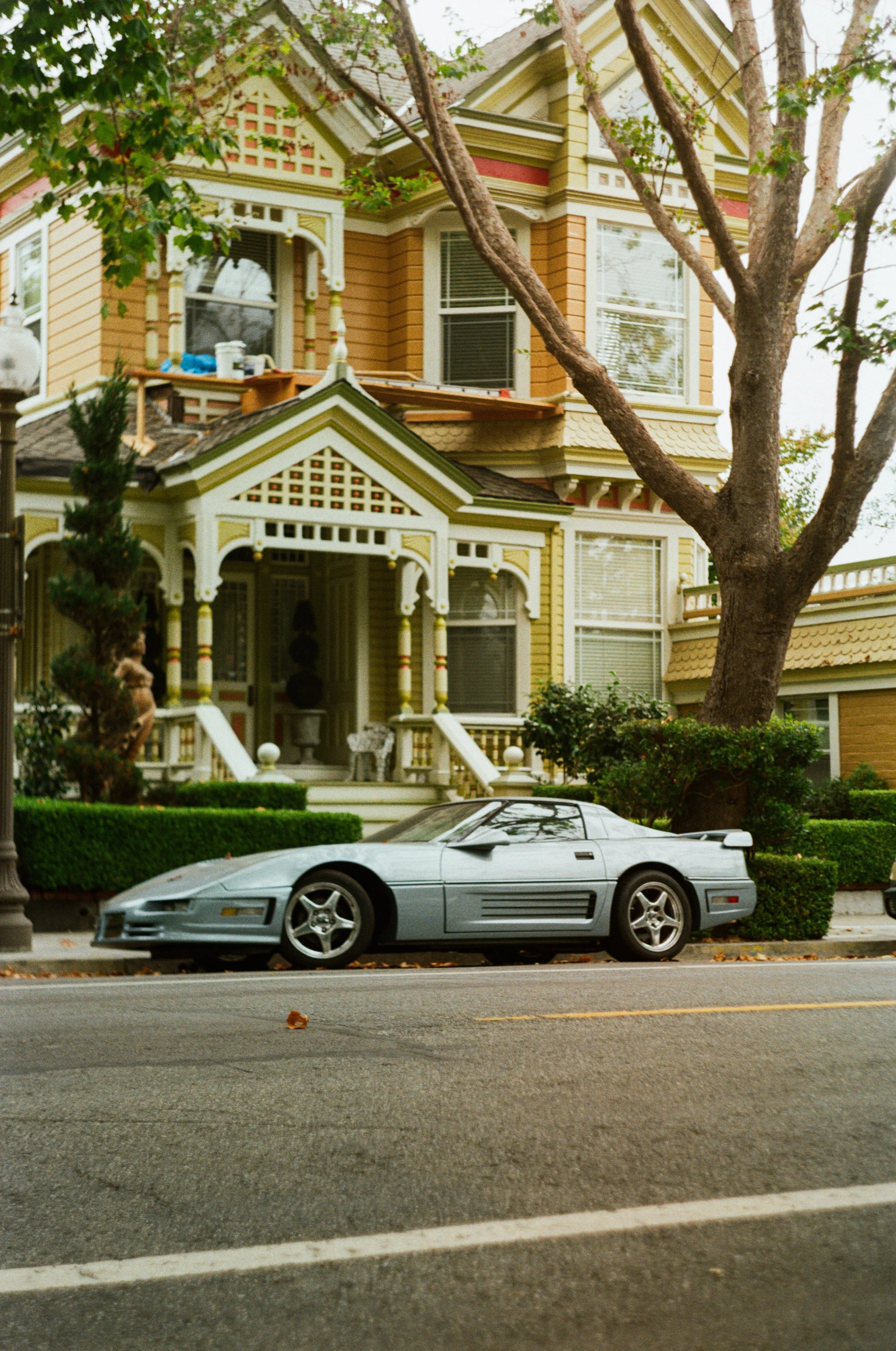 A silver sports car parked on the street in front of a large, ornate house with Victorian-style architecture, a porch, and a tree in the yard. Photo by Vincent Prograce