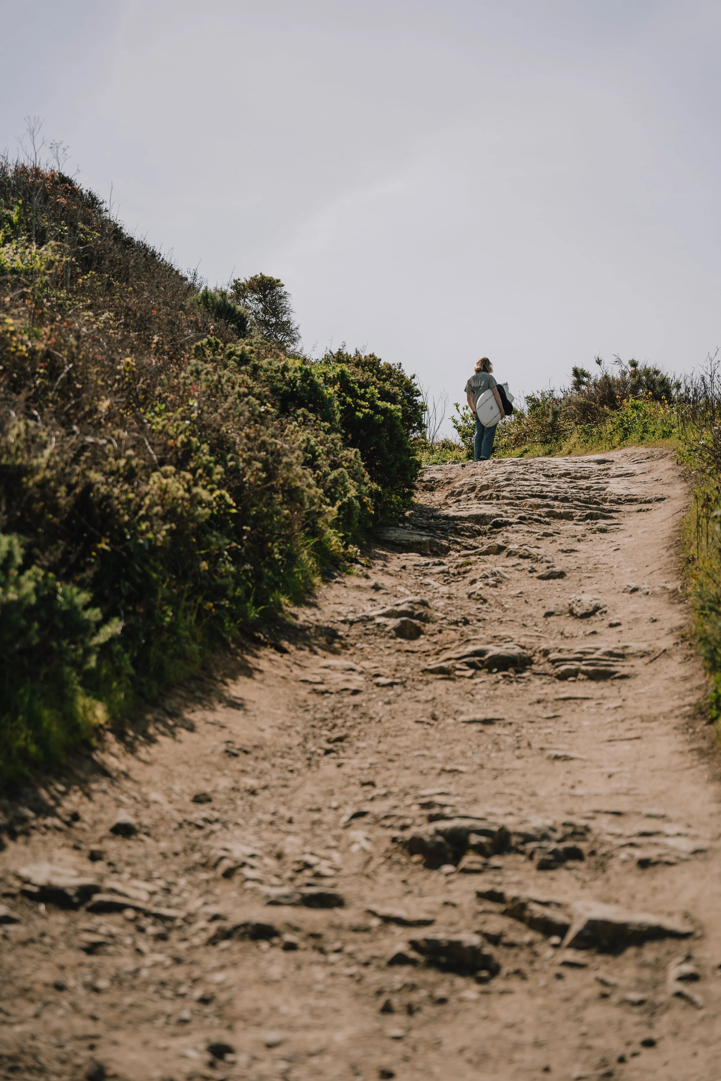 Person hiking on a rugged dirt trail surrounded by bushes and plants under a cloudy sky. Photo by Vincent Prograce