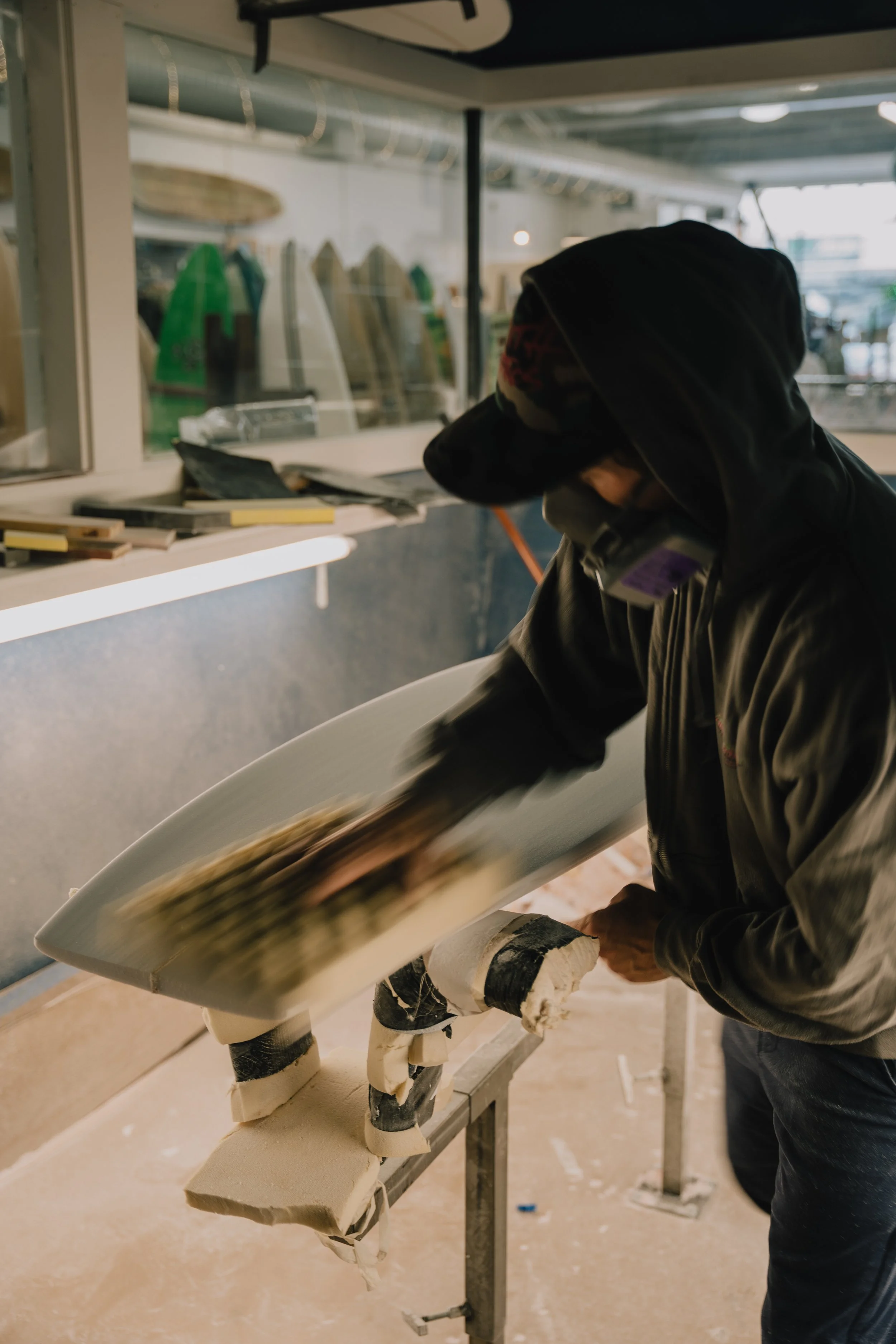 A person in a hoodie and face mask shaping a surfboard on a worktable in a workshop. Photo by Vincent Prograce