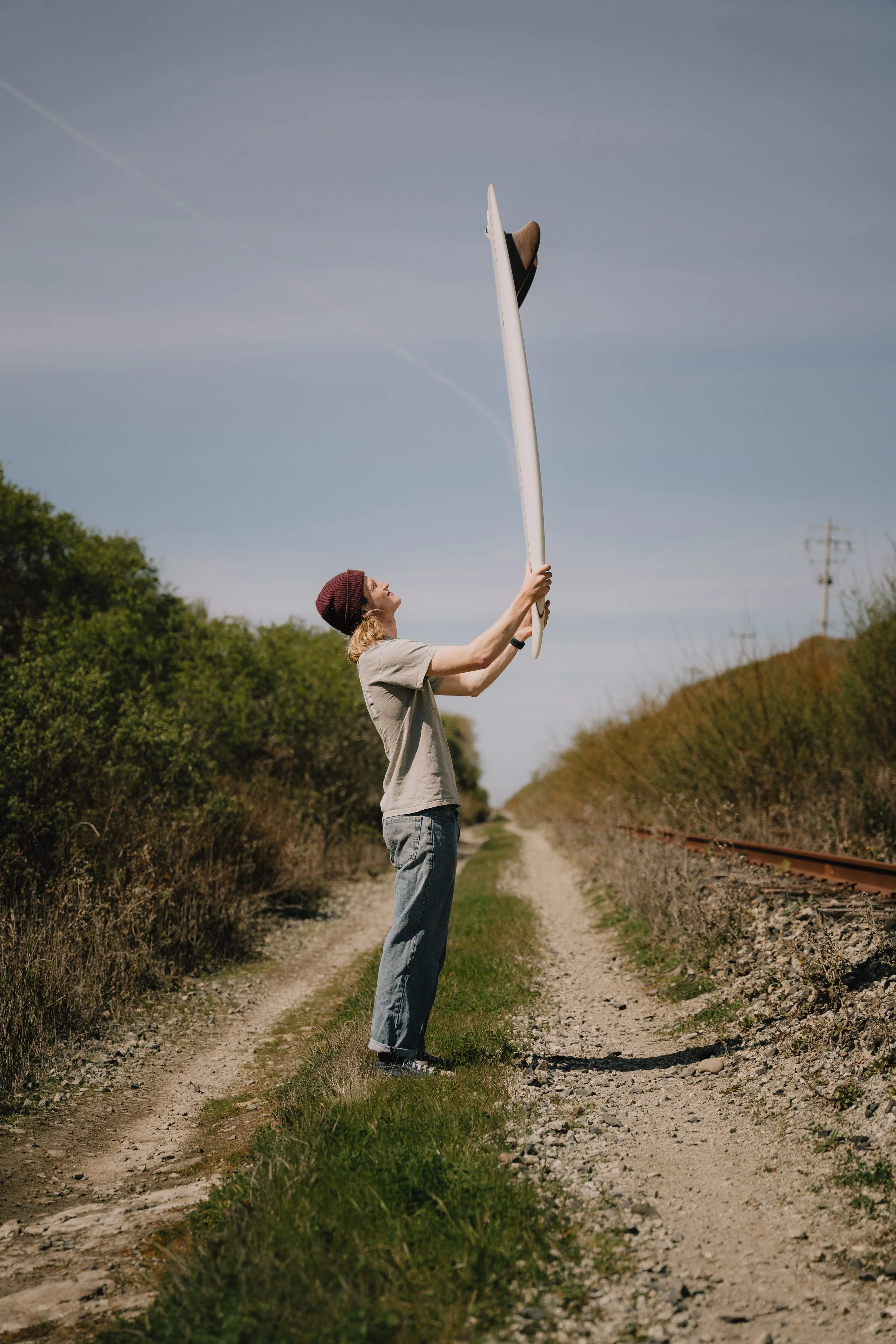 Person holding a surfboard above their head on a dirt path beside a railway track, with bushes and trees in the background under a clear sky. Photo by Vincent Prograce