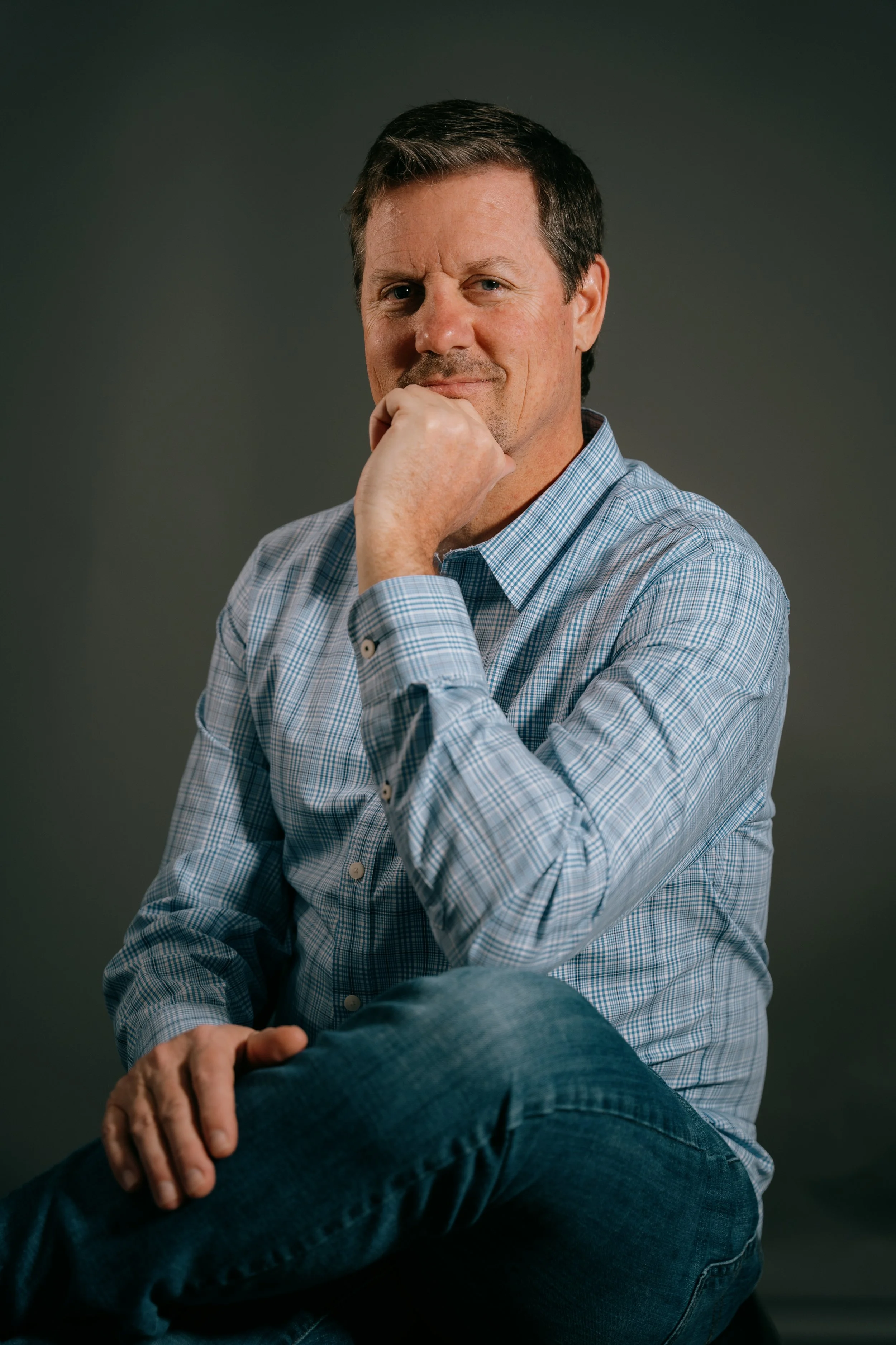Charlie Prograce a middle-aged man with dark hair, wearing a blue plaid shirt, sitting with one knee over the other, resting his chin on his fist with a thoughtful expression. Photo by Vincent Prograce