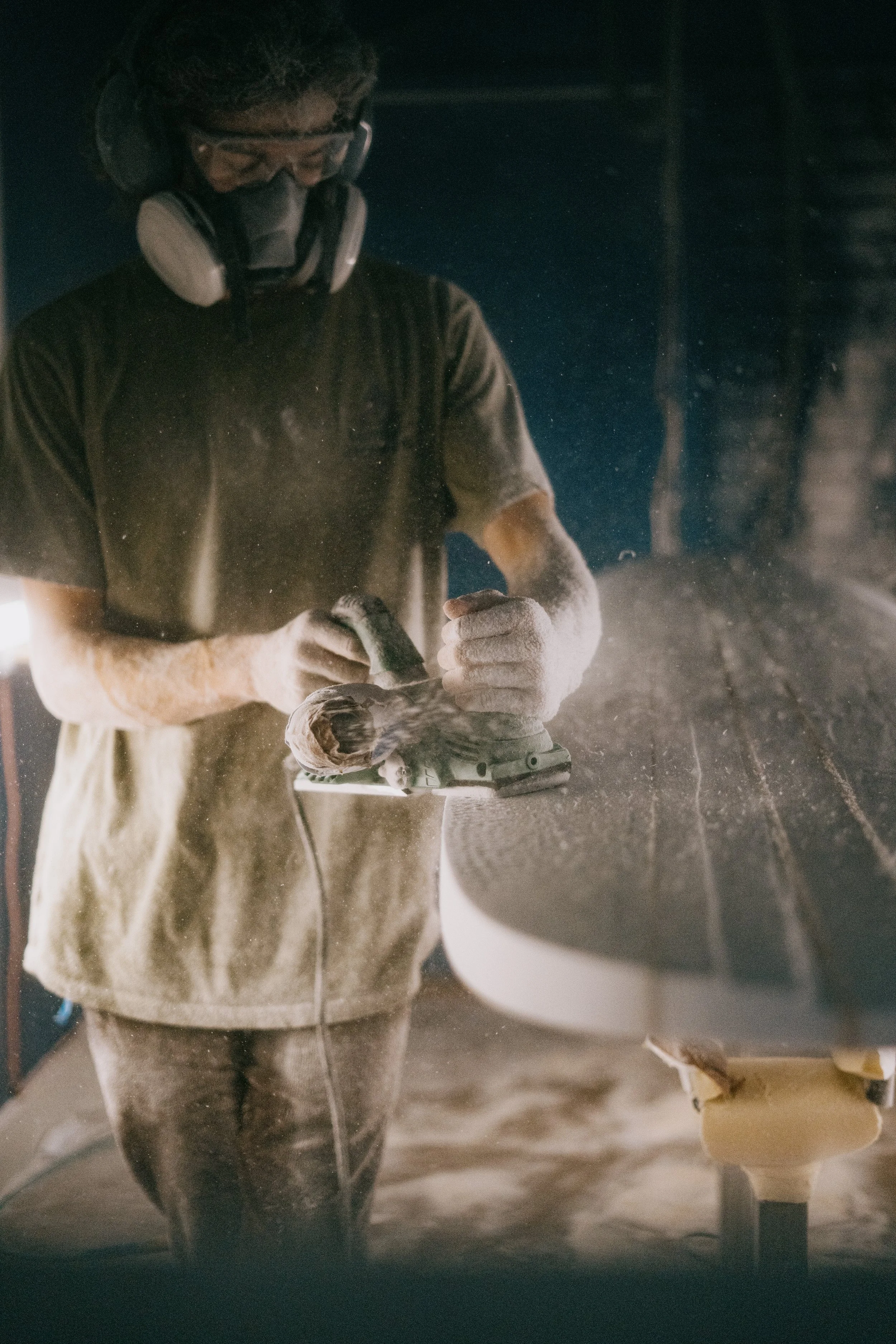 A person wearing safety goggles, a mask, and dust gloves is shaping a surfboard in a workshop filled with dust. Photo by Vincent Prograce