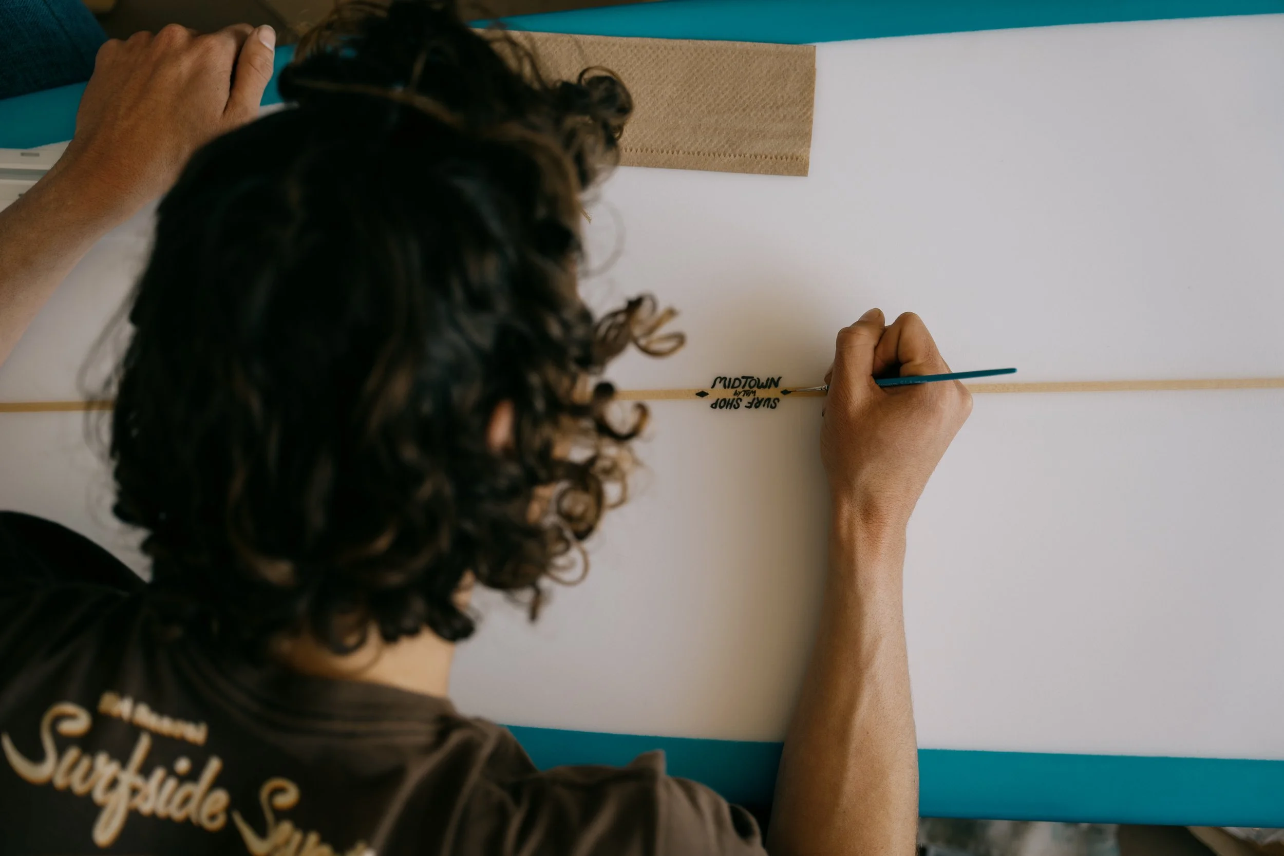 Person writing a sign on a surfboard for a surf shop, with the words 'Surf Shop Midtown' visible. Photo by Vincent Prograce