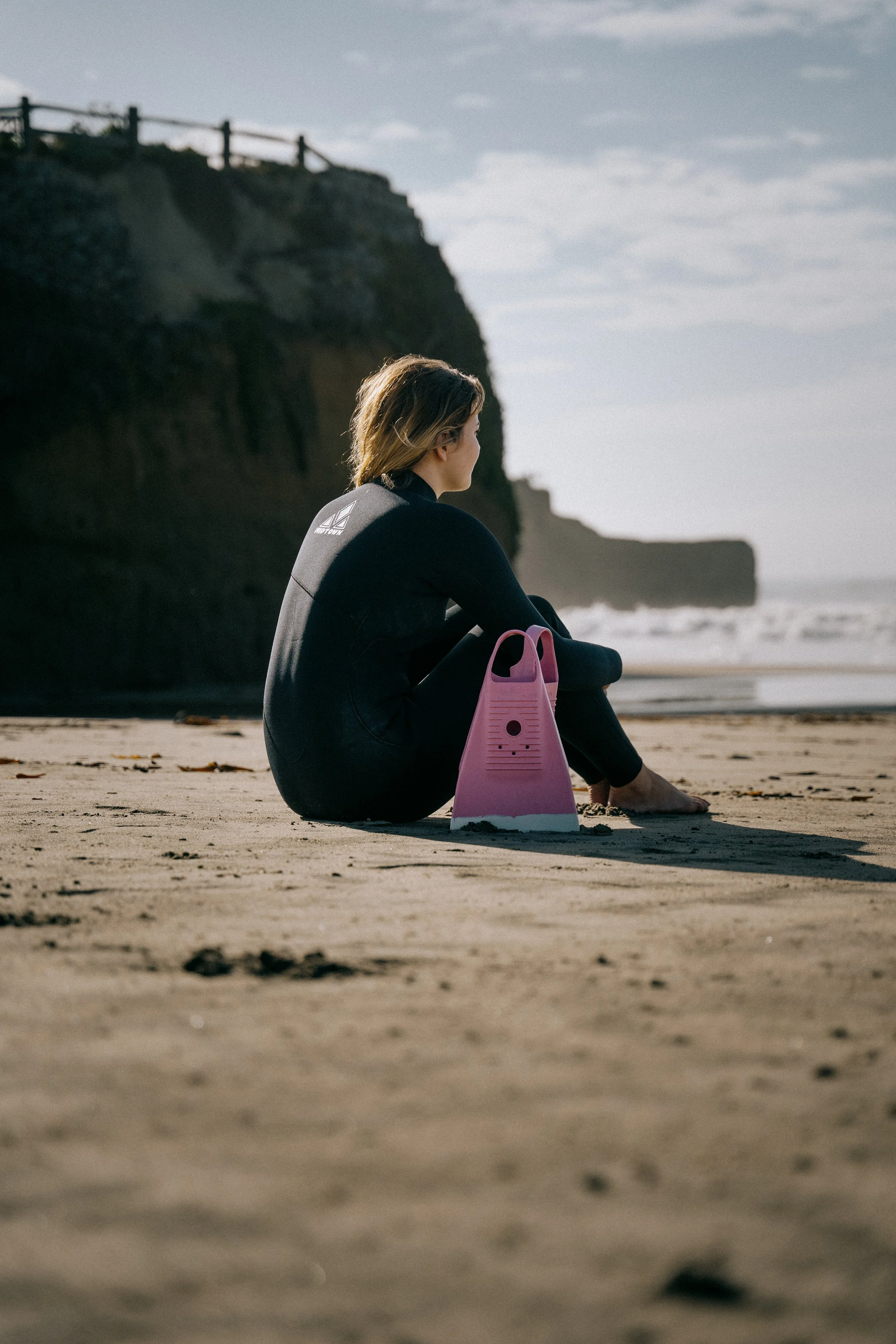 A woman in a black wetsuit sitting on a sandy beach next to a pink surfboard, looking towards the ocean with cliffs in the background. Photo by Vincent Prograce