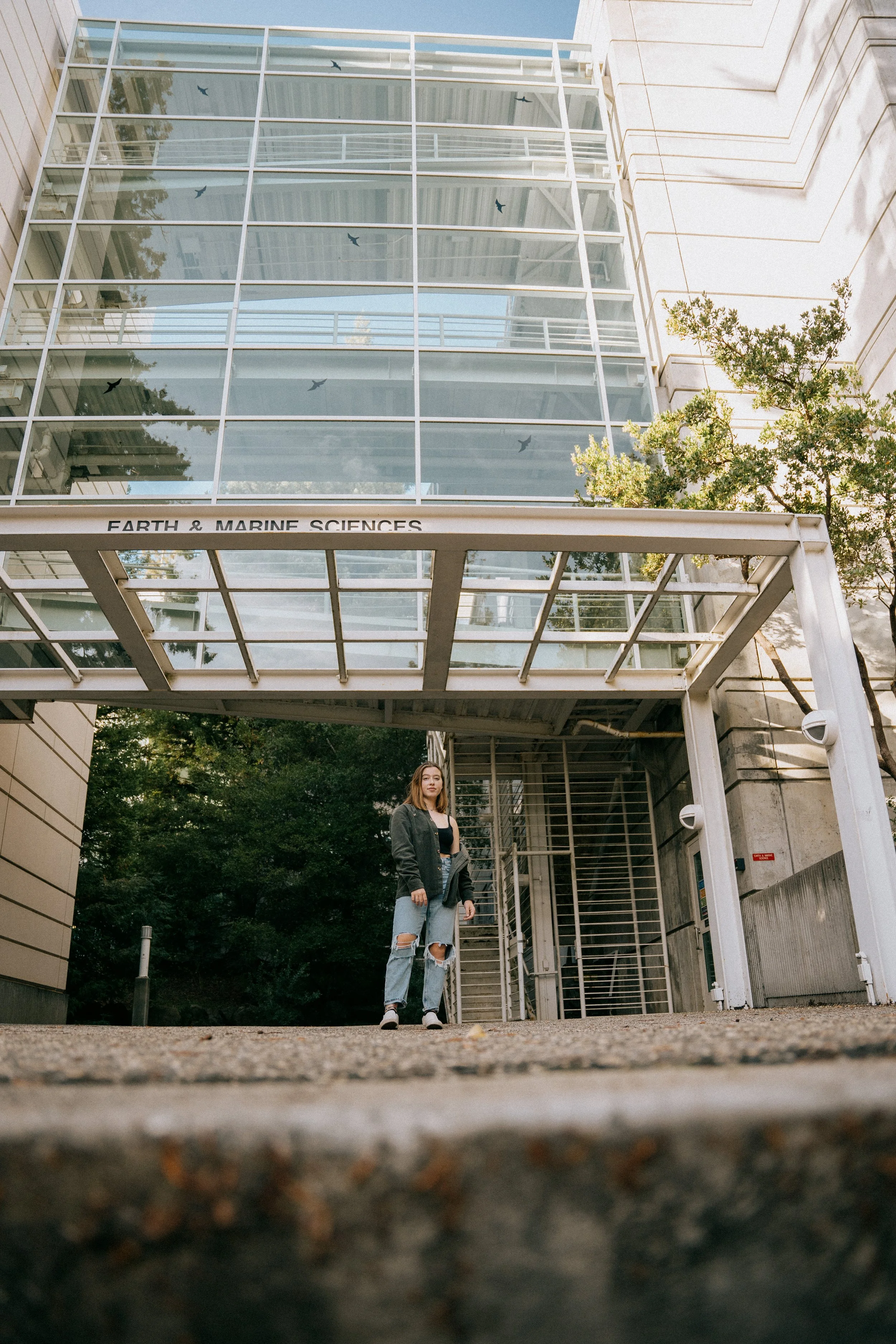 A young woman stands outside the Earth & Marine Sciences building, which has a modern glass and steel architectural design, with trees and a partly cloudy sky in the background. Photo by Vincent Prograce