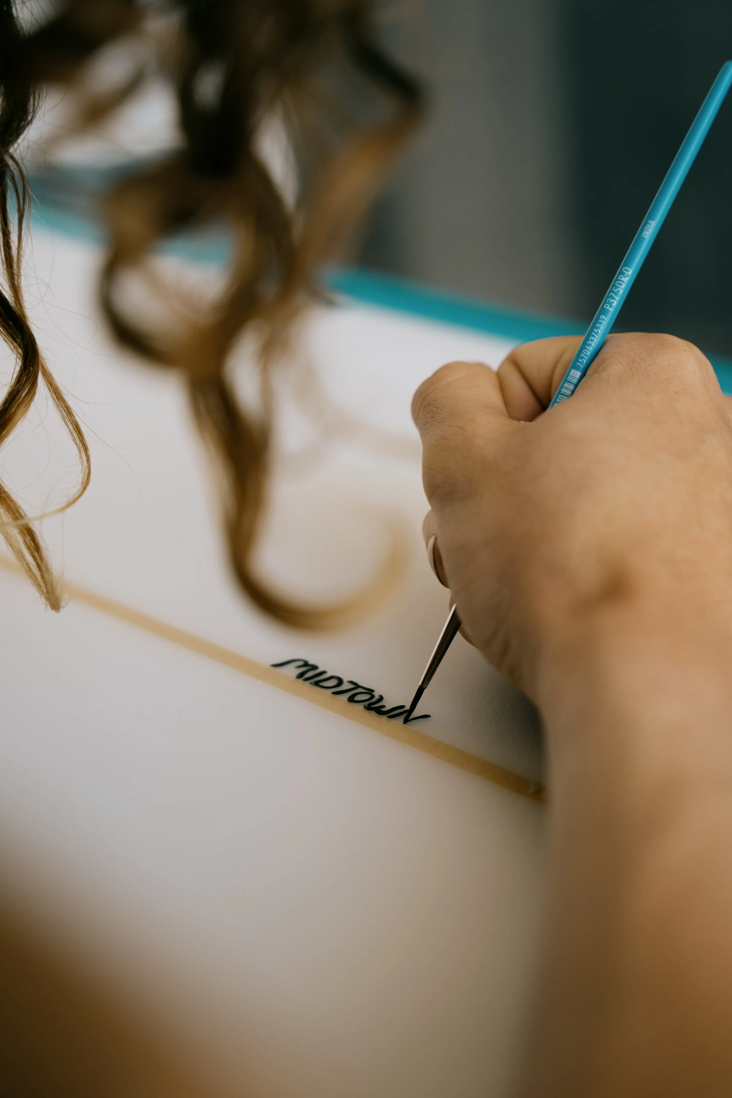 A person is writing in cursive on a white surfboard with a paint brush; their curly hair is partially visible on the left side of the image. Photo by Vincent Prograce