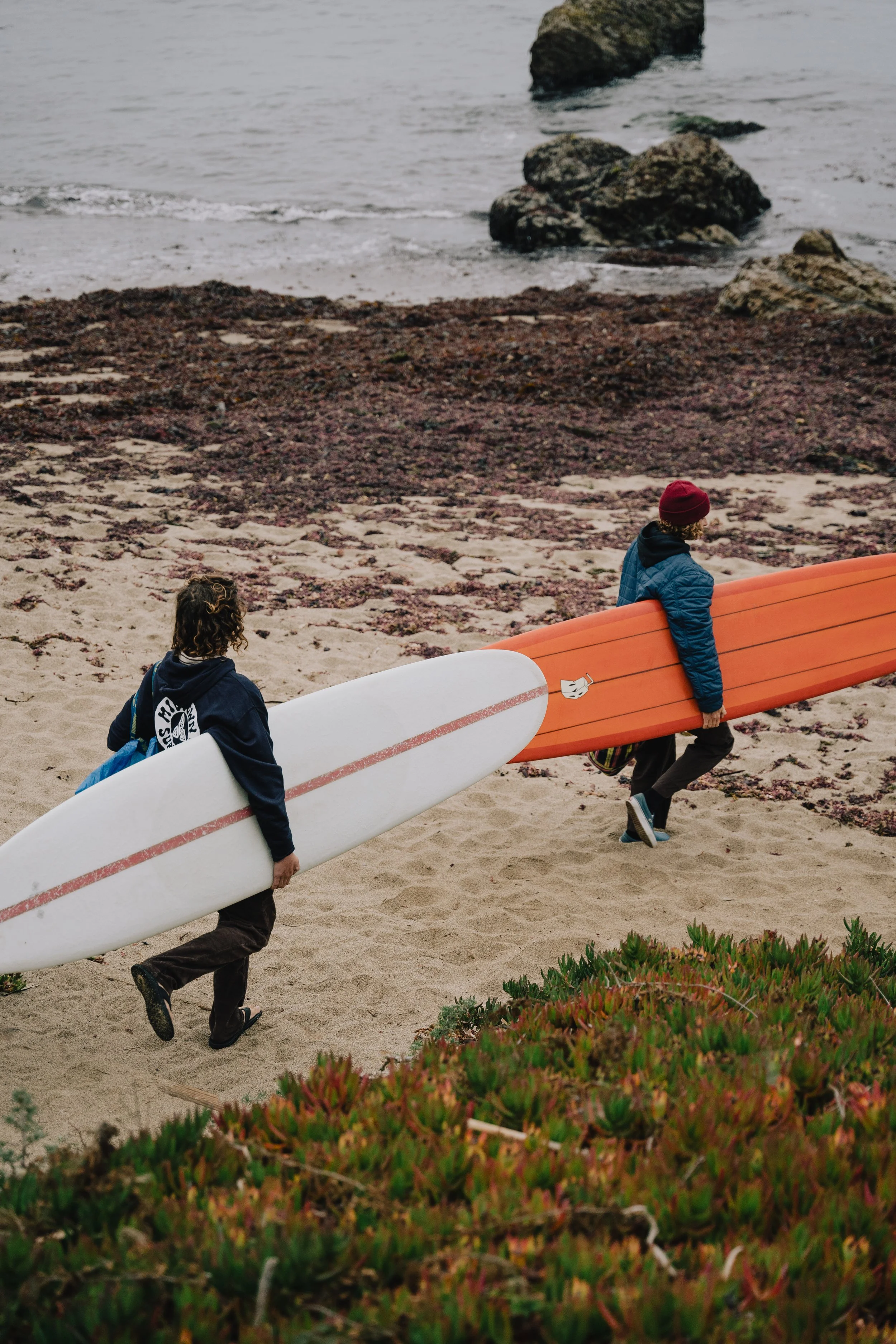 Two children carrying surfboards on a sandy beach, walking toward the ocean, with rocks in the background. Photo by Vincent Prograce