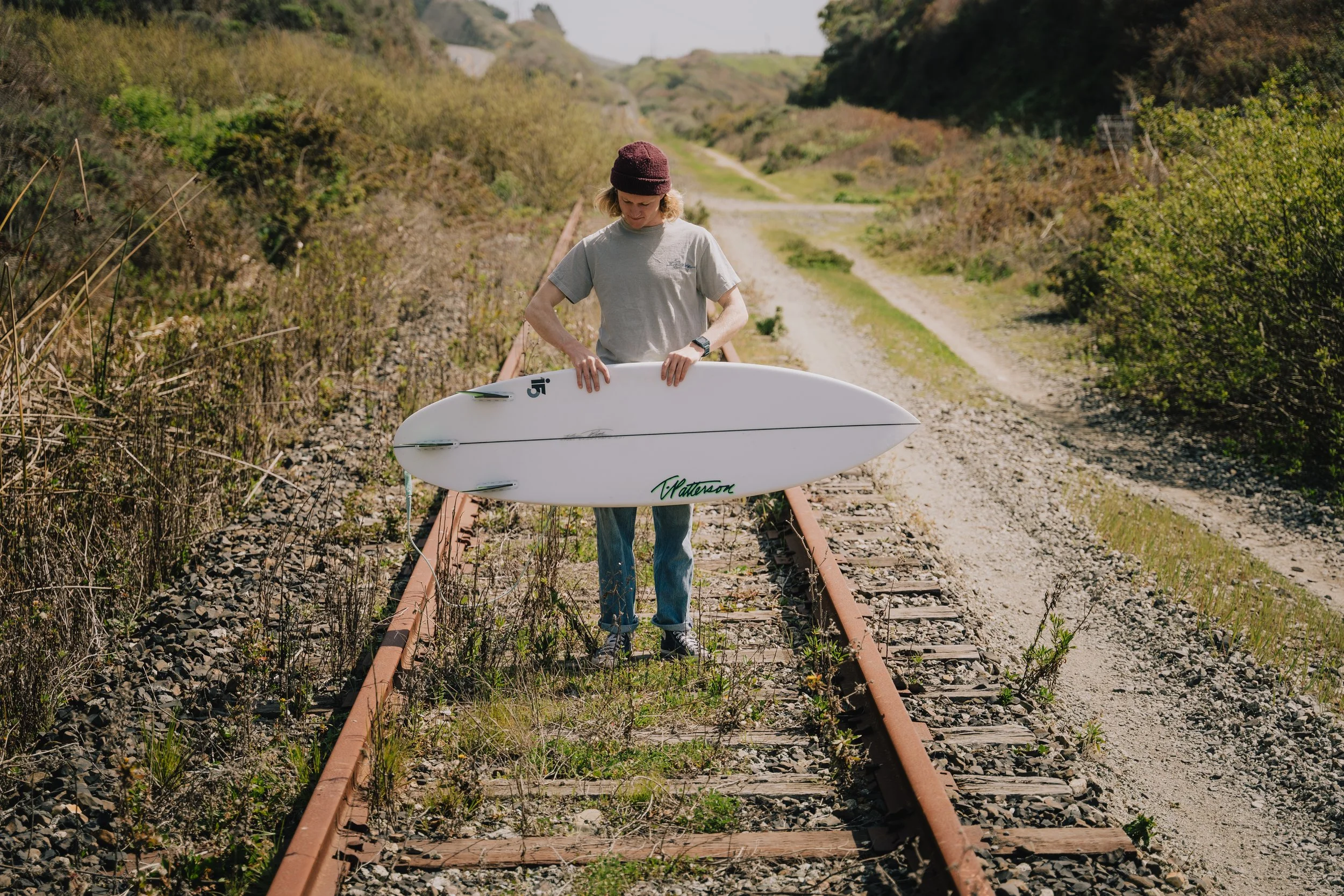A person standing on an abandoned train track holding a white surfboard, with greenery and a dirt path in the background. Photo by Vincent Prograce