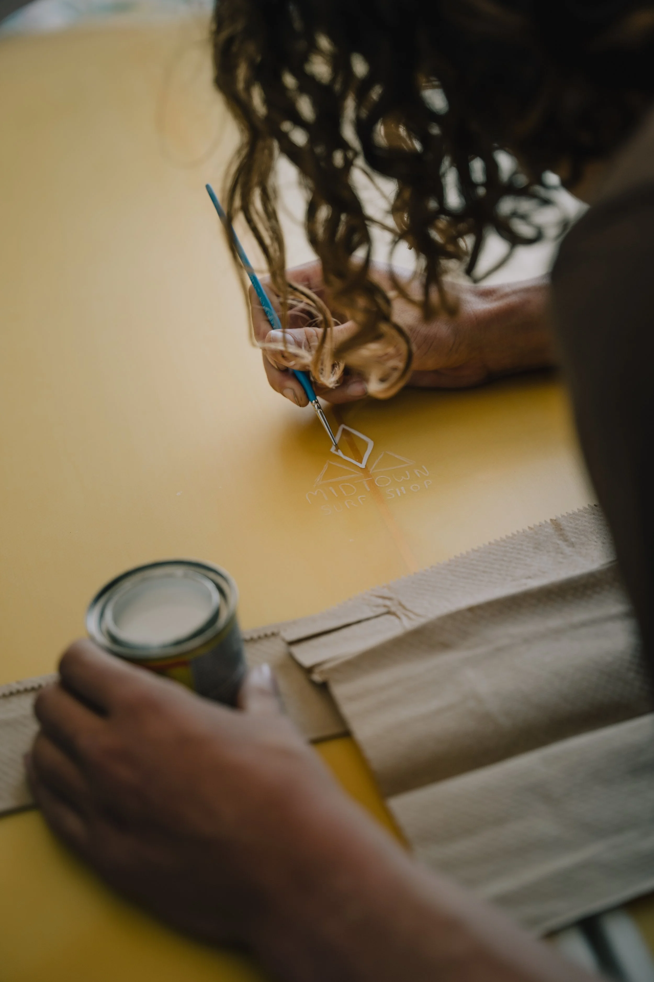 A person with curly hair is drawing a logo for Midtown Surf Shop on a yellow surfboard with white chalk, using a blue paintbrush, while holding a small container of white paint. Photo by Vincent Prograce