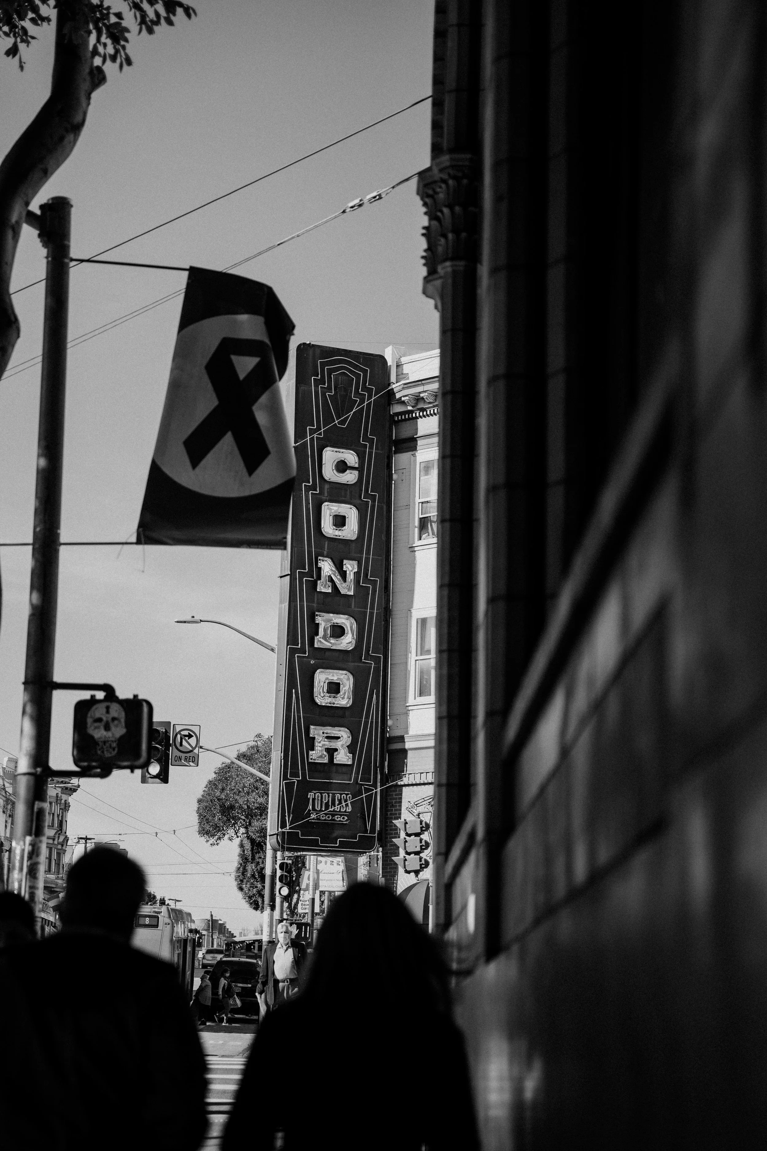 Black and white photo of a San Francisco city street scene with a vertical sign reading 'CONDER' in large letters, pedestrians walking, and a flag with a ribbon icon hanging from a pole. Photo by Vincent Prograce
