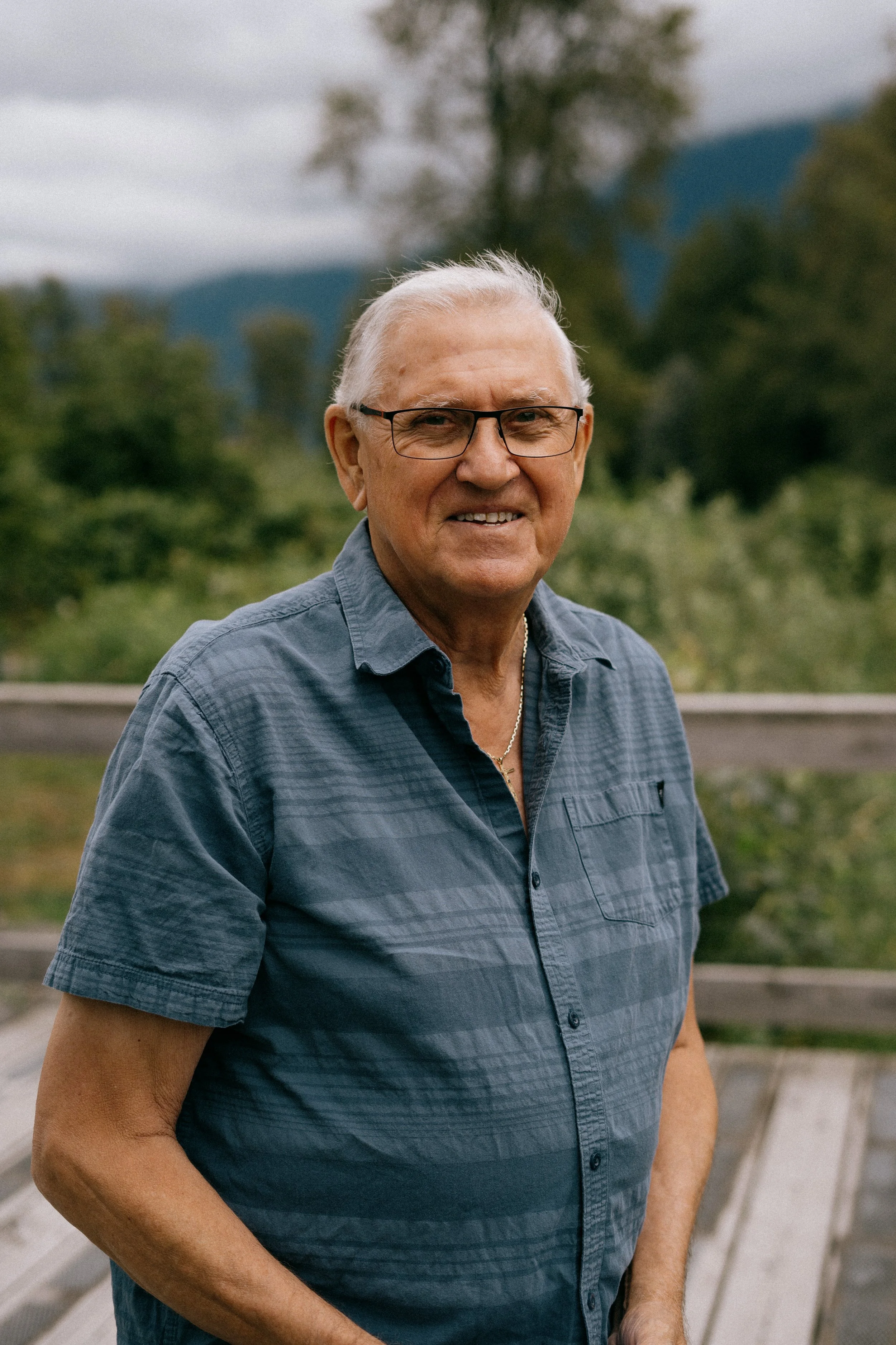 An elderly man with glasses and gray hair, wearing a blue short-sleeved shirt, standing outdoors with a mountain and forested landscape in the background. Photo by Vincent Prograce