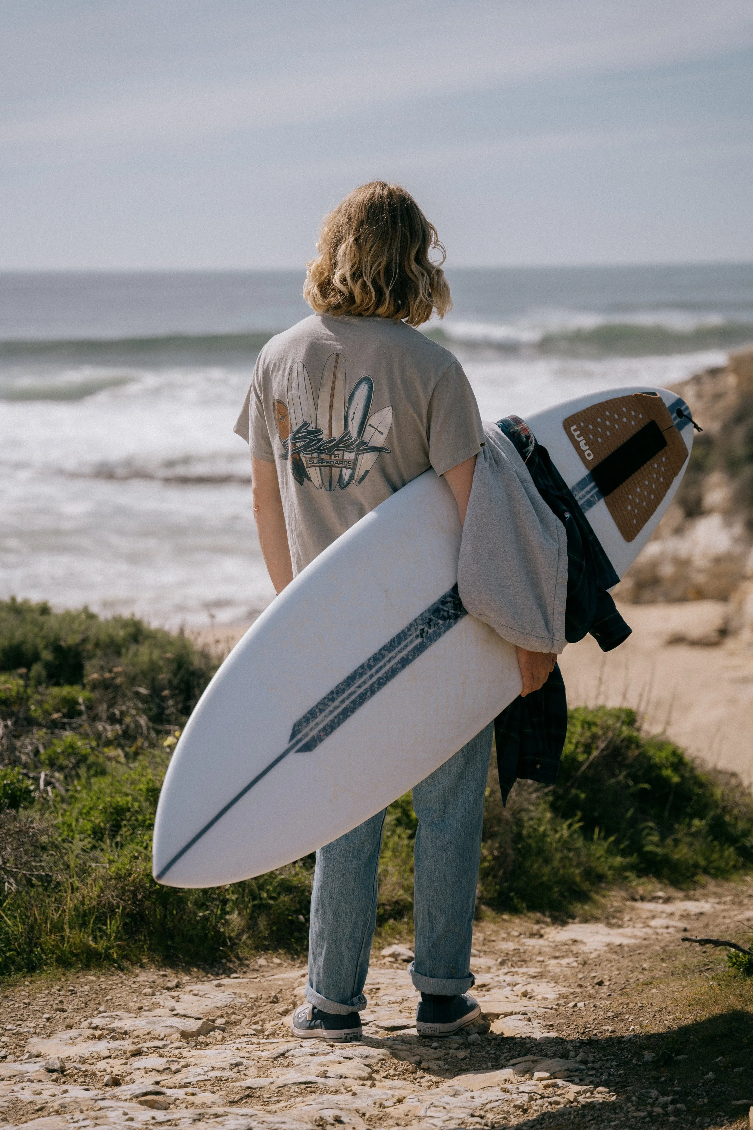 A person with shoulder-length wavy hair stands on a rocky path near the beach, holding a surfboard under their arm and looking at the ocean. Photo by Vincent Prograce