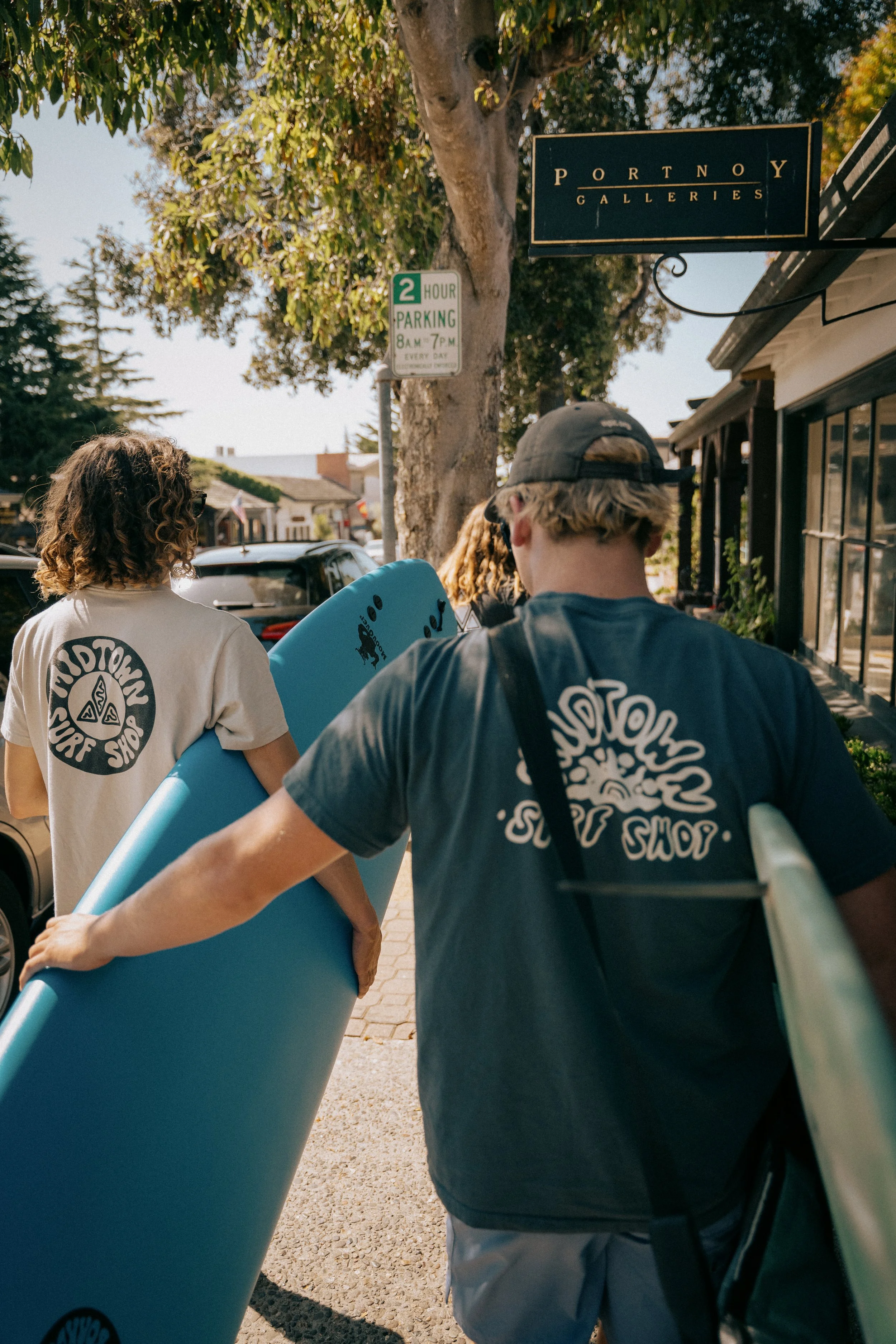 People carrying surfboards walk on a sidewalk in front of a store. Photo by Vincent Prograce