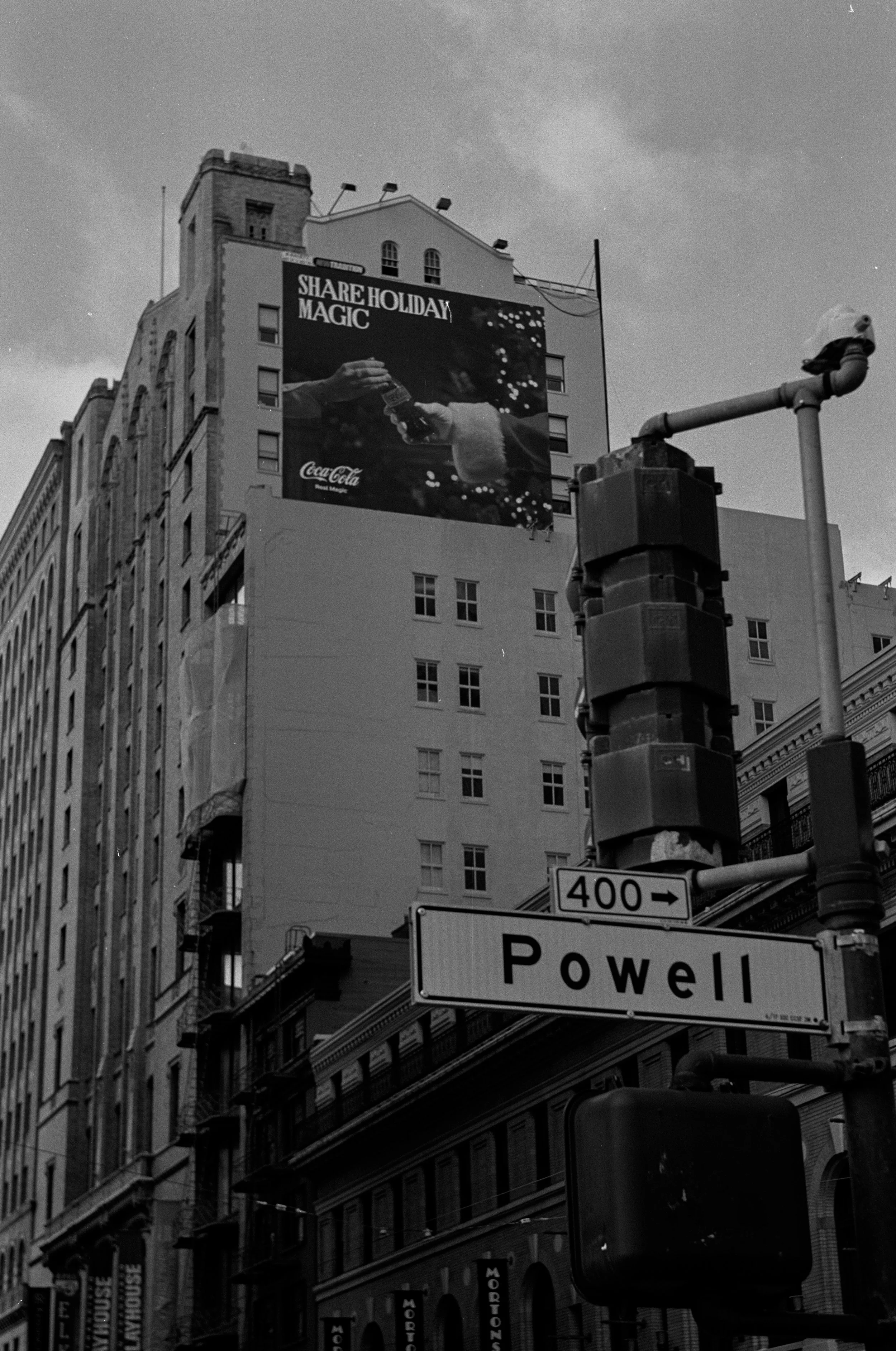 city street scene with a tall building featuring a Coca-Cola advertisement, a street sign reading 'Powell,' and a traffic signal. The advertisement shows a person pouring Coca-Cola with the text 'Share Holiday Magic.' Photo by Vincent Prograce
