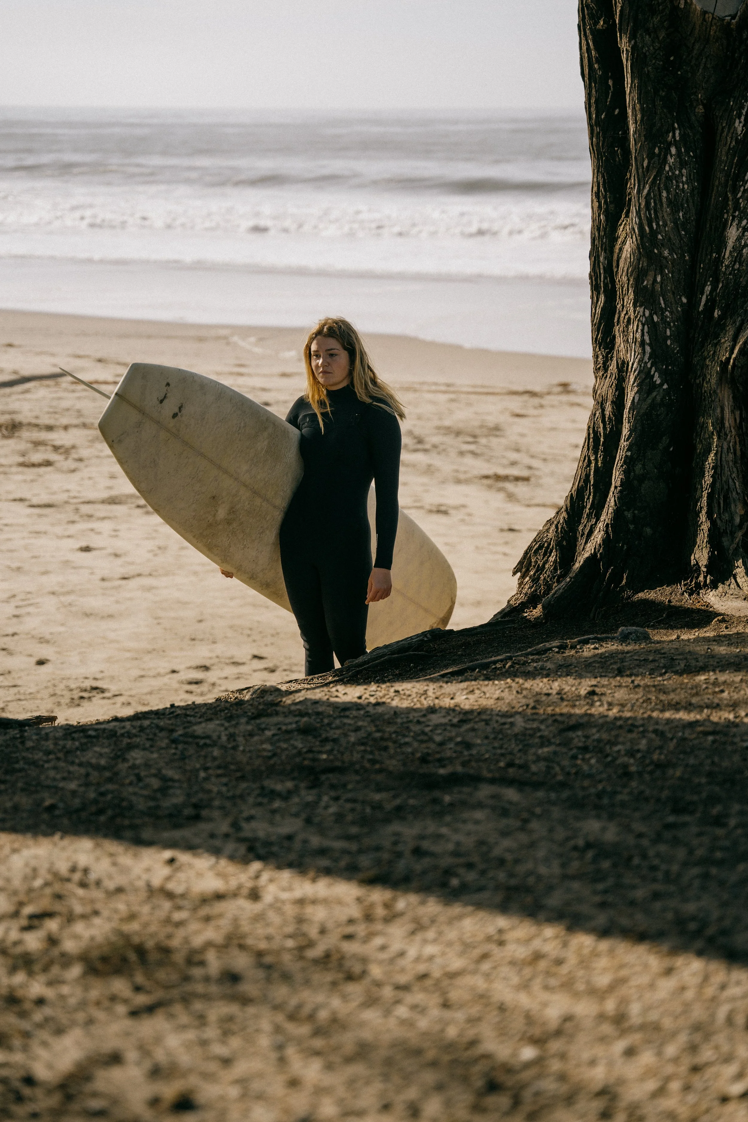 A woman dressed in a black wetsuit standing on a sandy beach holding a surfboard under her arm near a large tree, with the ocean waves in the background. Photo by Vincent Prograce
