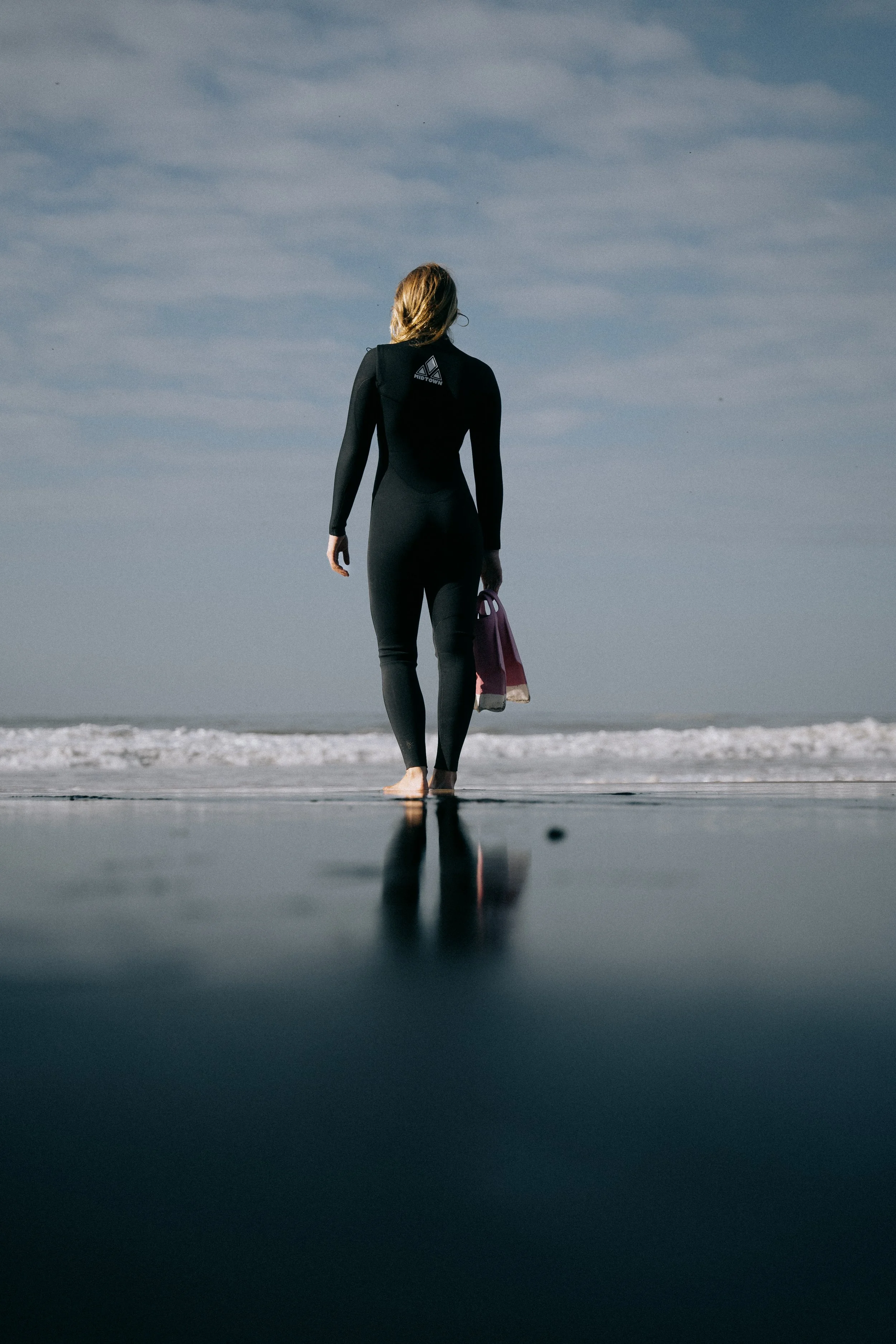 A woman in a black wetsuit walking barefoot on the beach holding a swim fins, with ocean waves and cloudy sky in the background. Photo by Vincent Prograce
