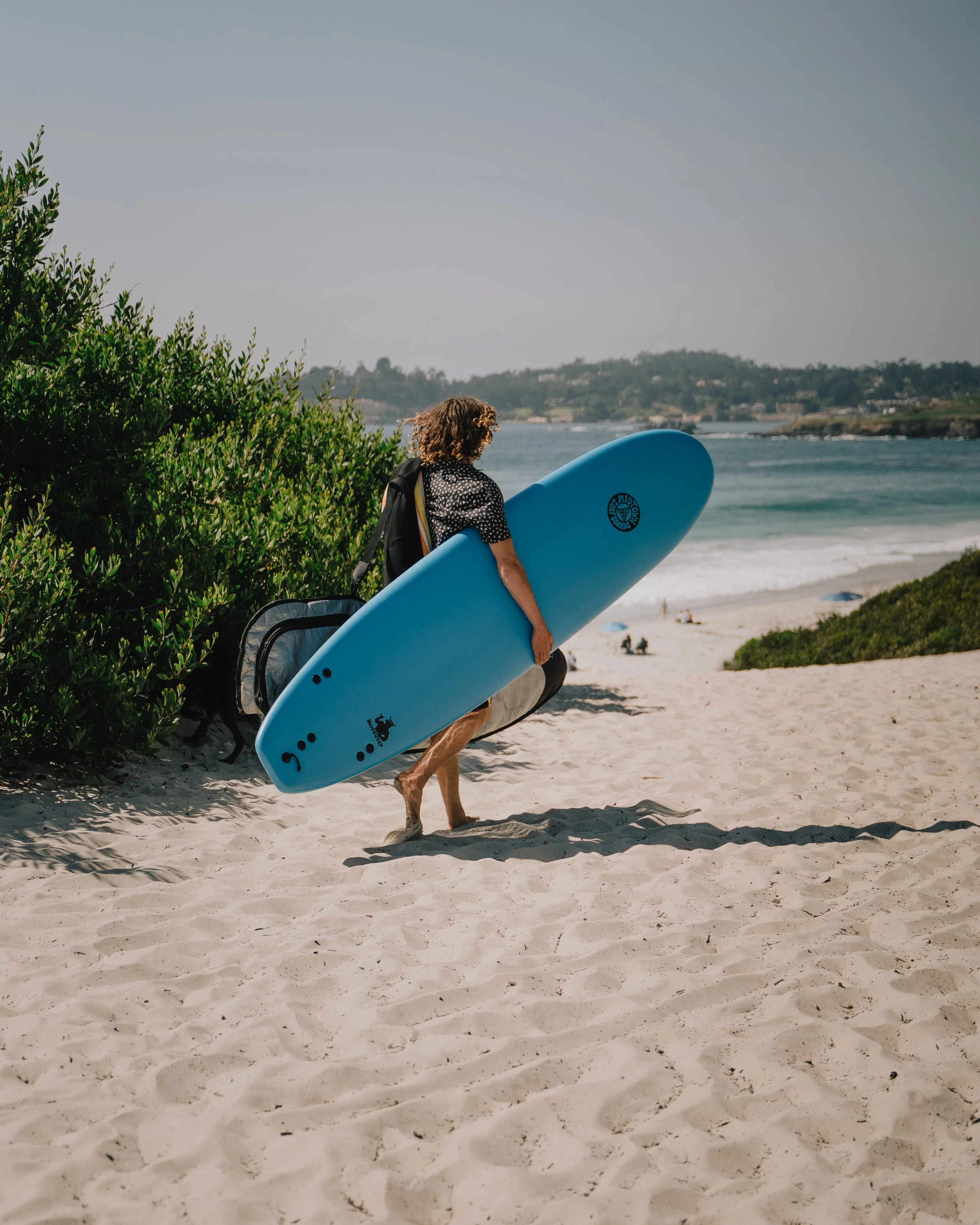 Person walking on beach carrying a blue surfboard with a backpack. Photo by Vincent Prograce