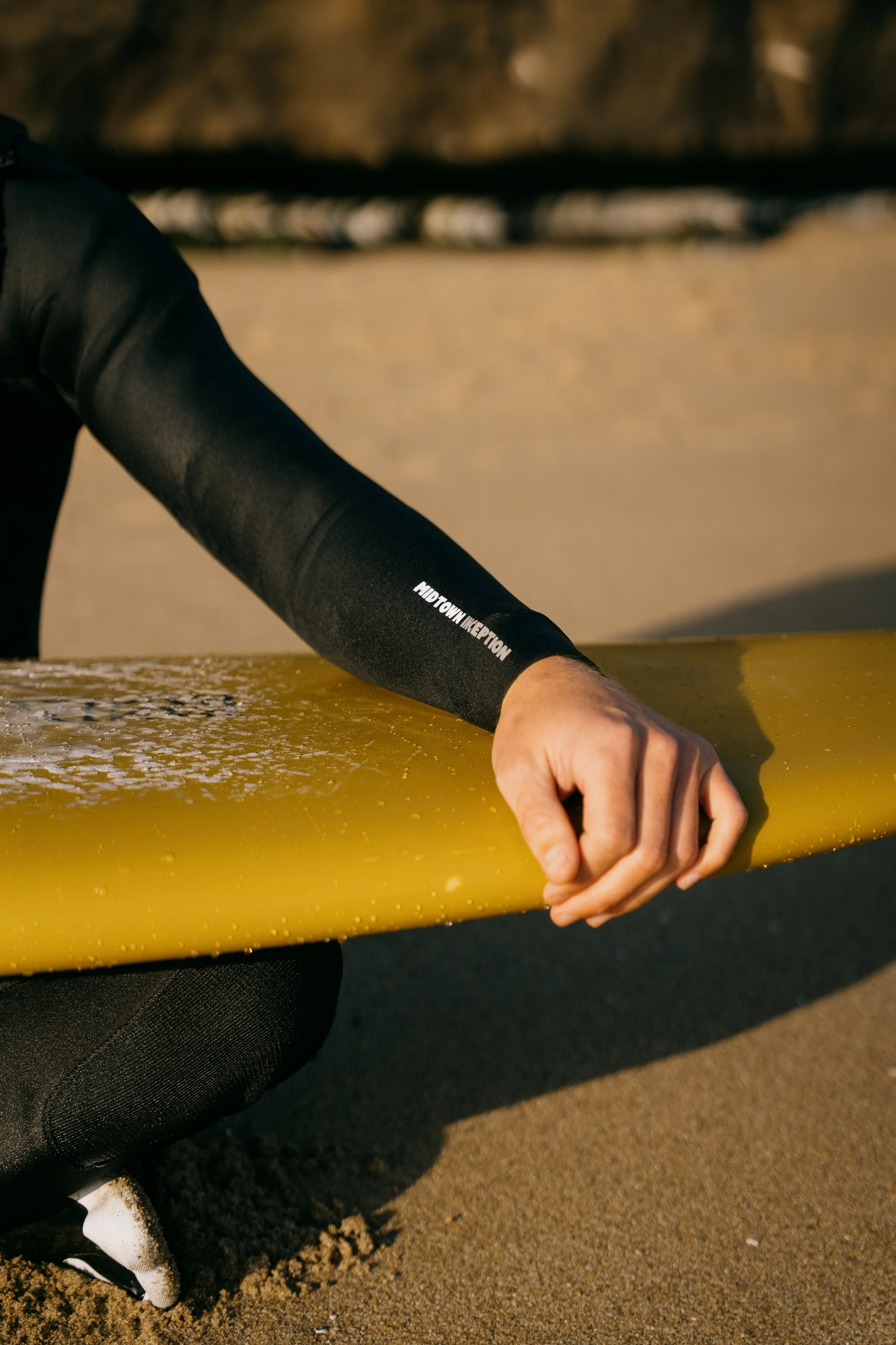 Surfer in wetsuit sitting on the sand next to a yellow surfboard with water droplets, at the beach. Photo by Vincent Prograce