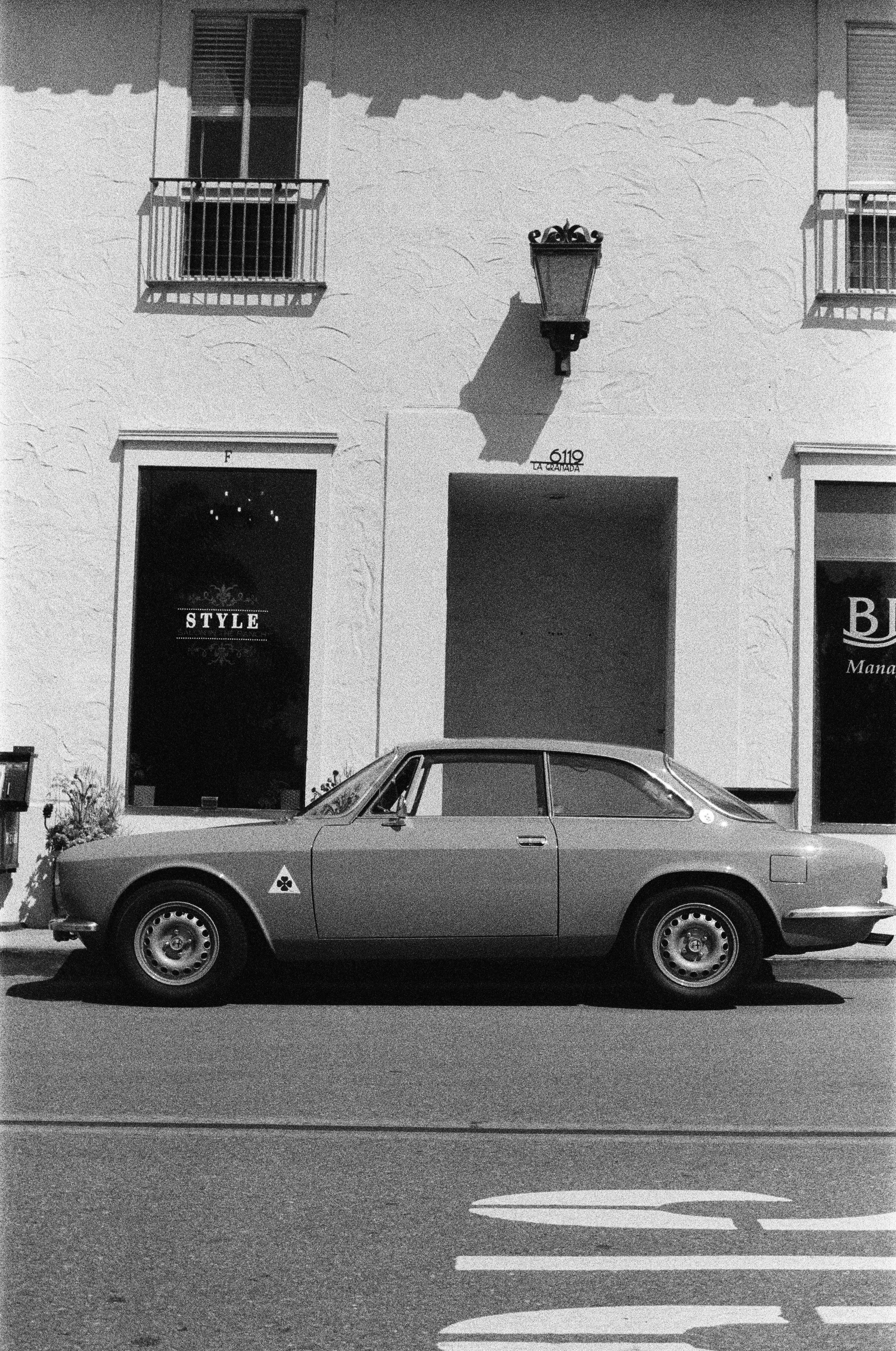 A black and white photo of a vintage car parked on the street in front of a building with balconies, a lantern, and signs reading 'STYLE' and 'BE'. Photo by Vincent Prograce