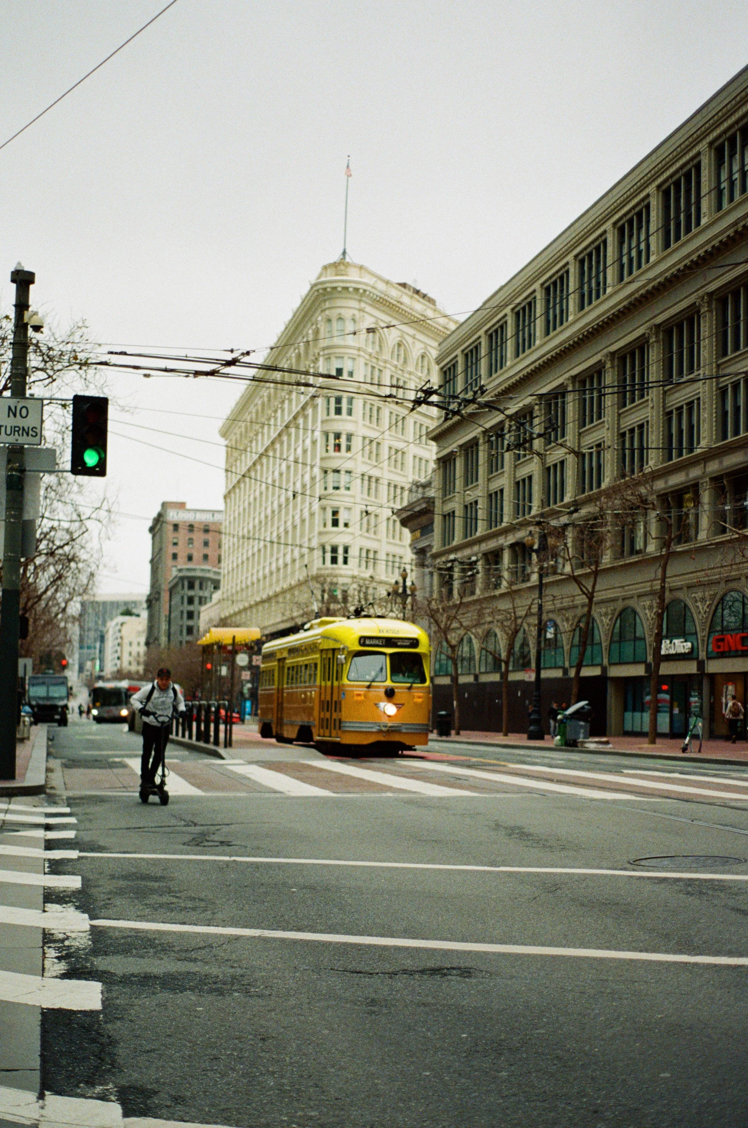 A yellow streetcar traveling down a city street in San Francisco, California, with pedestrians walking and buildings lining the street. Photo by Vincent Prograce