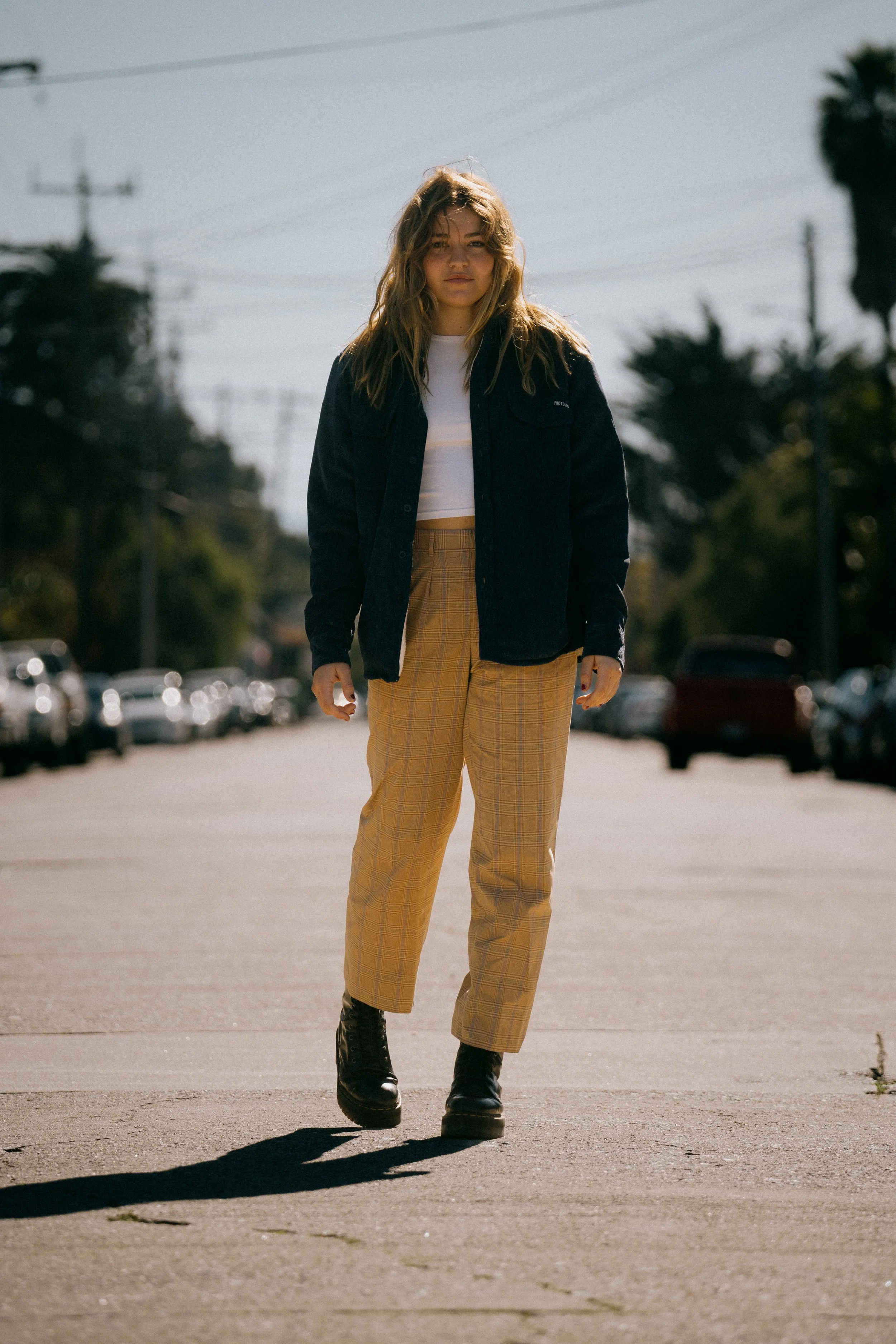 A young woman walking on a city street, wearing a black jacket, white t-shirt, yellow plaid pants, and black boots, with trees and parked cars in the background. Photo by Vincent Prograce