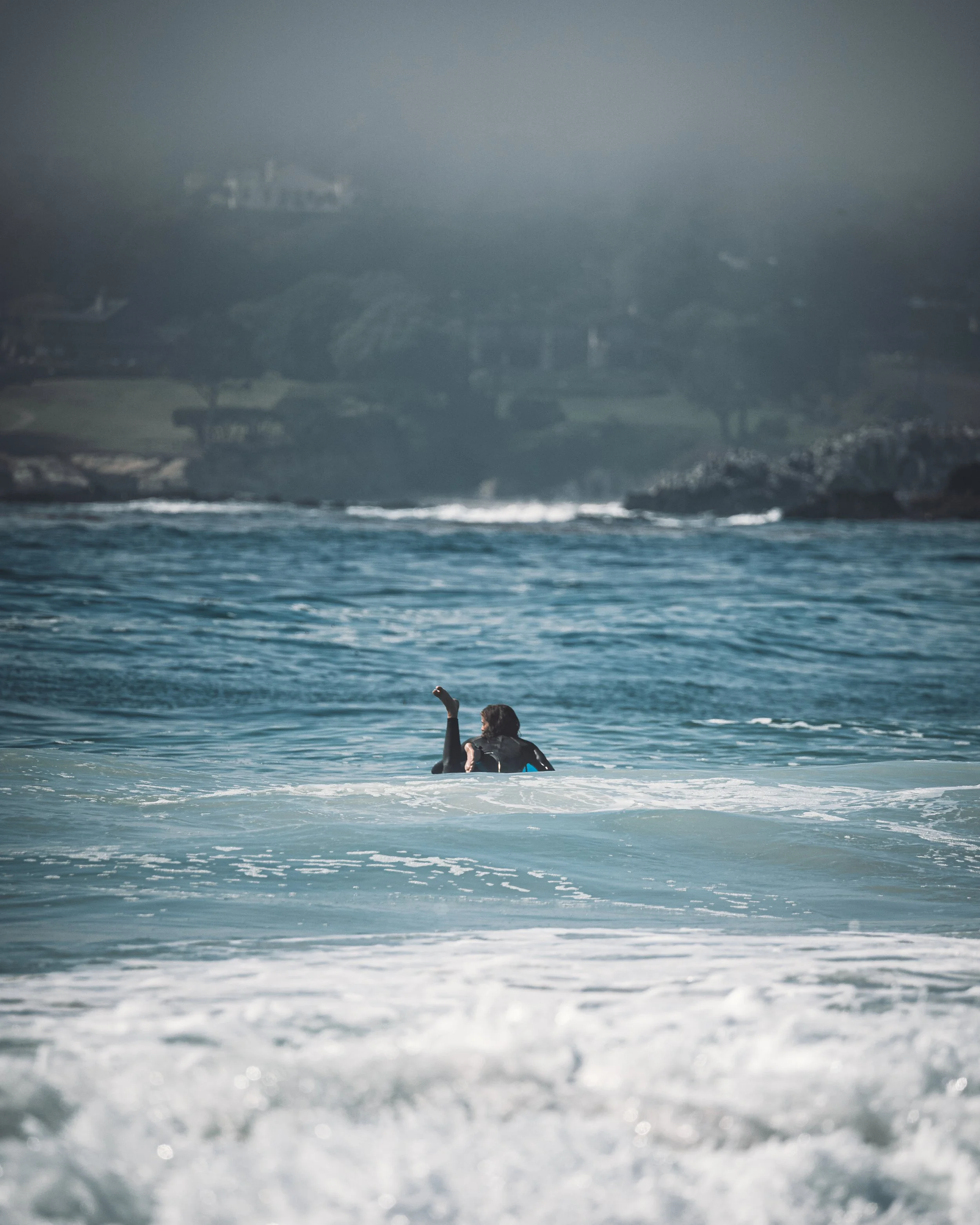 A surfer in the ocean, facing away, with land and houses visible in the distance. Photo by Vincent Prograce