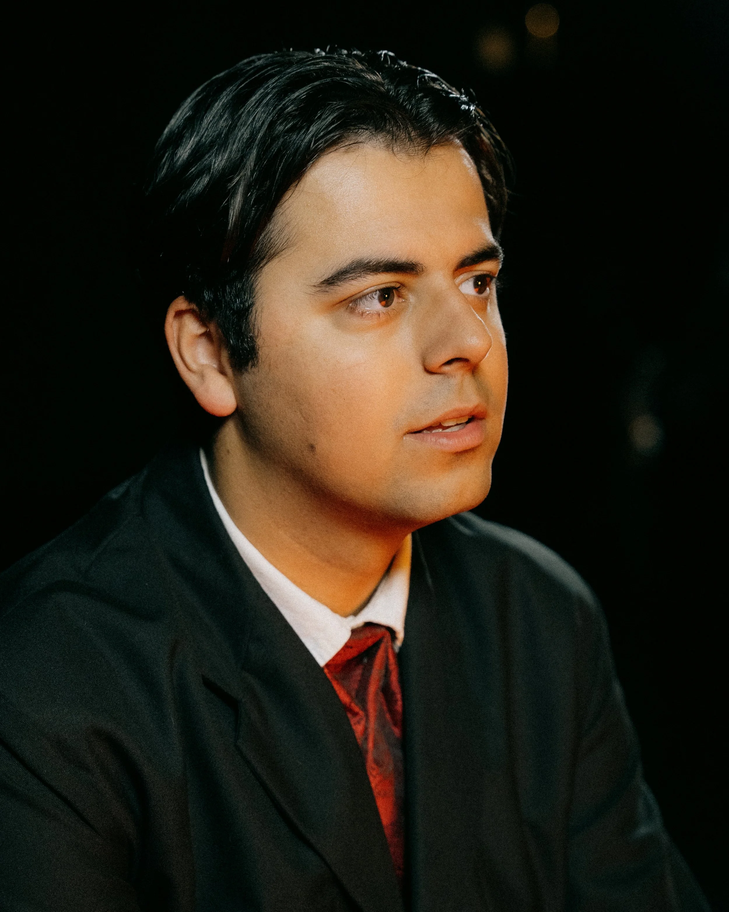 A young man with dark hair and light skin, wearing a black suit, white shirt, and red tie, looking to his right against a dark background. Photo by Vincent Prograce