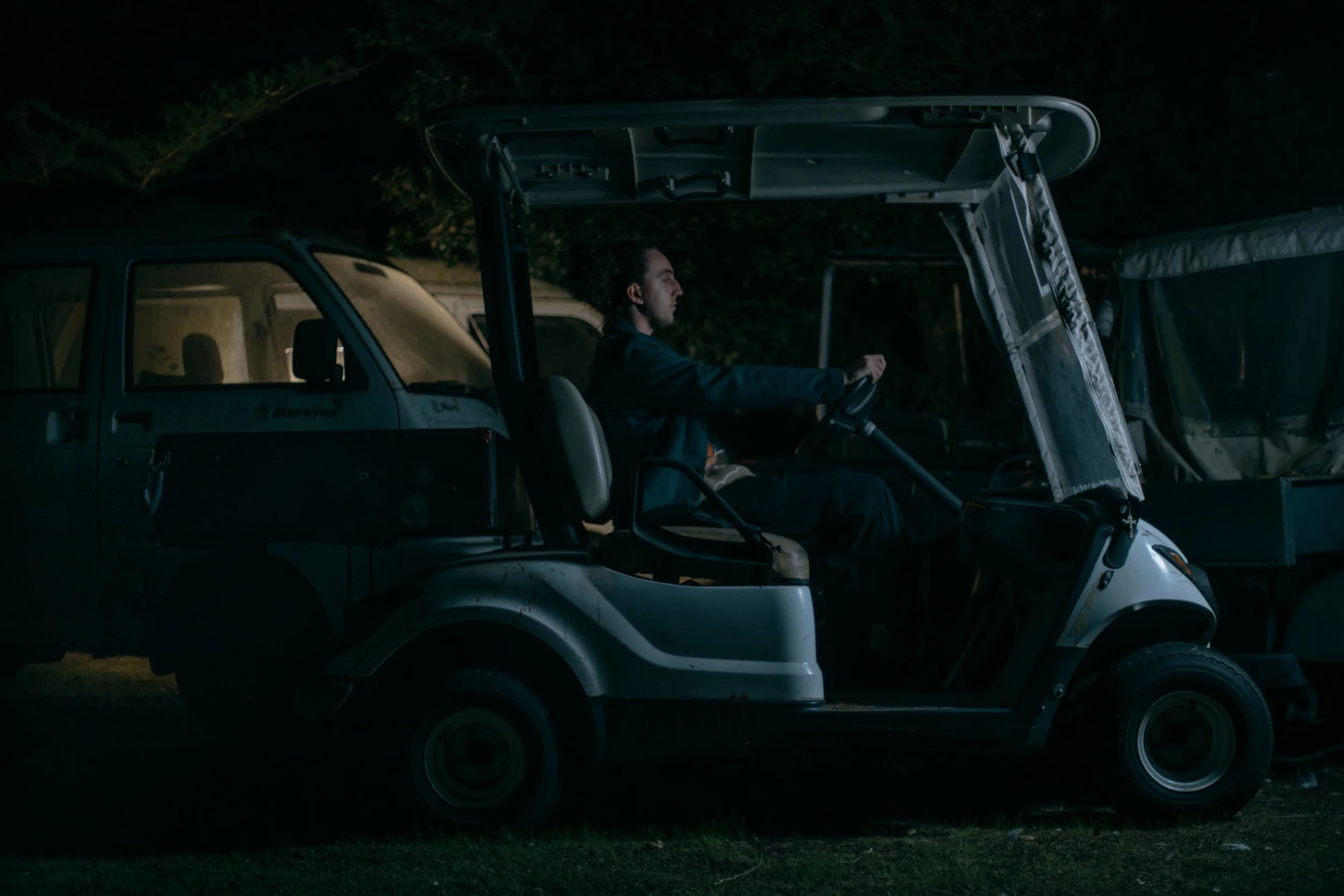 A man driving a golf cart at night, surrounded by parked utility vehicles, with trees in the background. Photo by Vincent Prograce