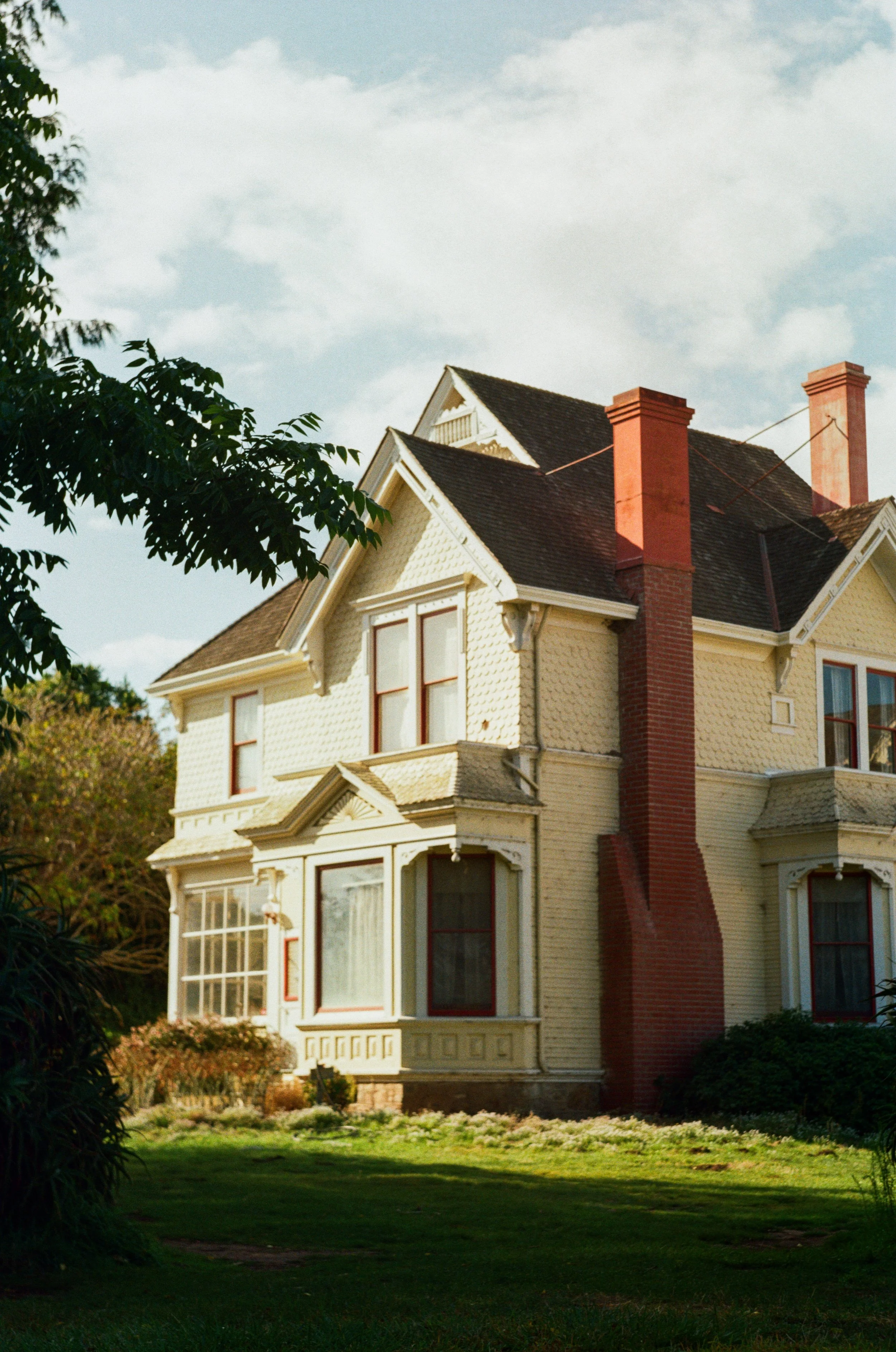 A Victorian-style house with yellow paint, a large red brick chimney, and tall windows, surrounded by a well-maintained lawn and trees under a partly cloudy sky. Photo by Vincent Prograce
