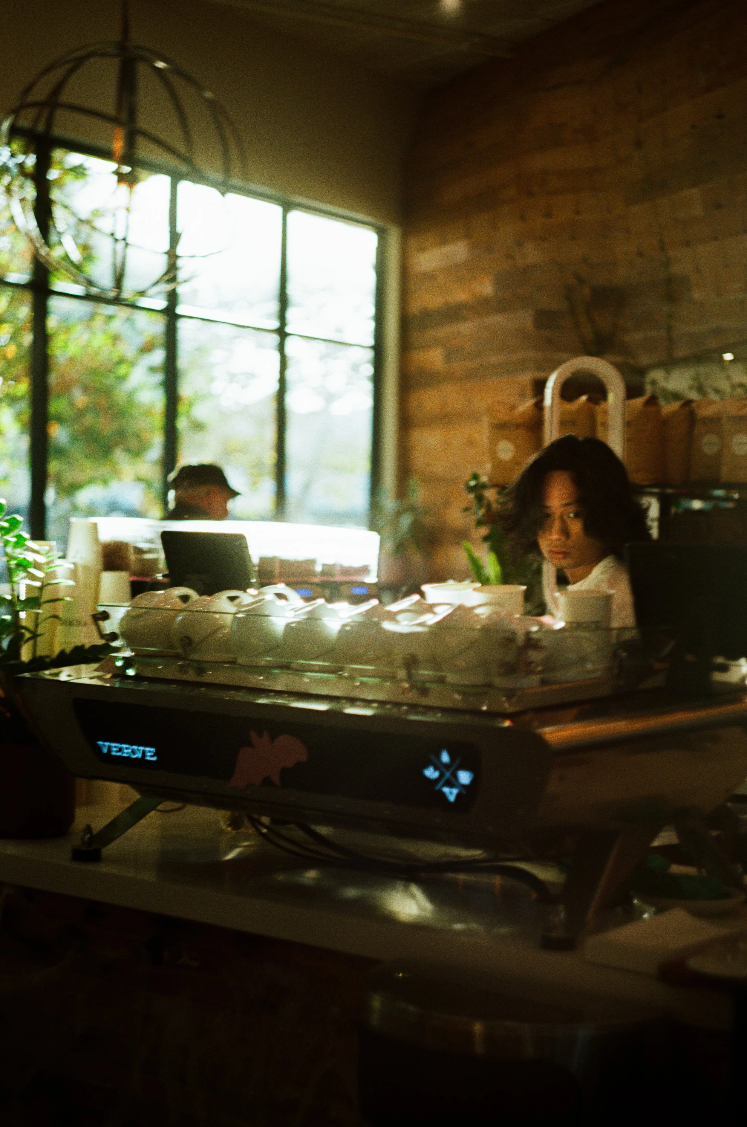 A man with shoulder-length dark hair working behind the counter of a coffee shop, with large windows in the background. Photo by Vincent Prograce