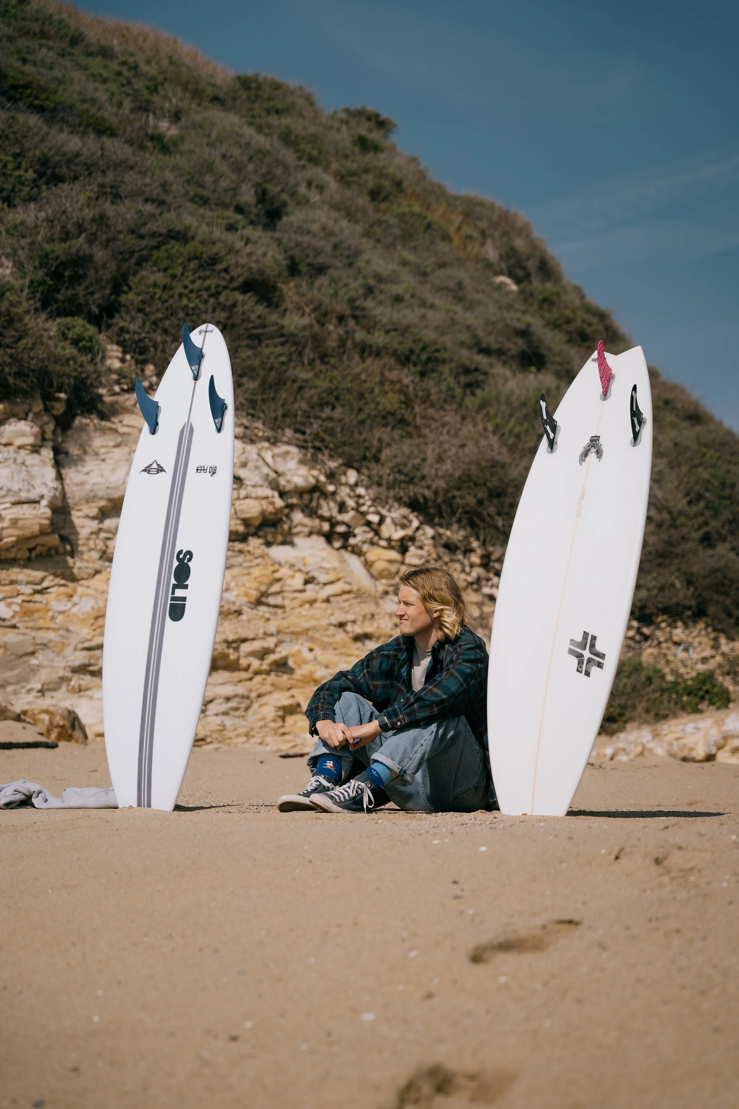 A person sitting on the sand at the beach, surrounded by two stand-up paddleboards, with rocky cliffs and green hills in the background. Photo by Vincent Prograce