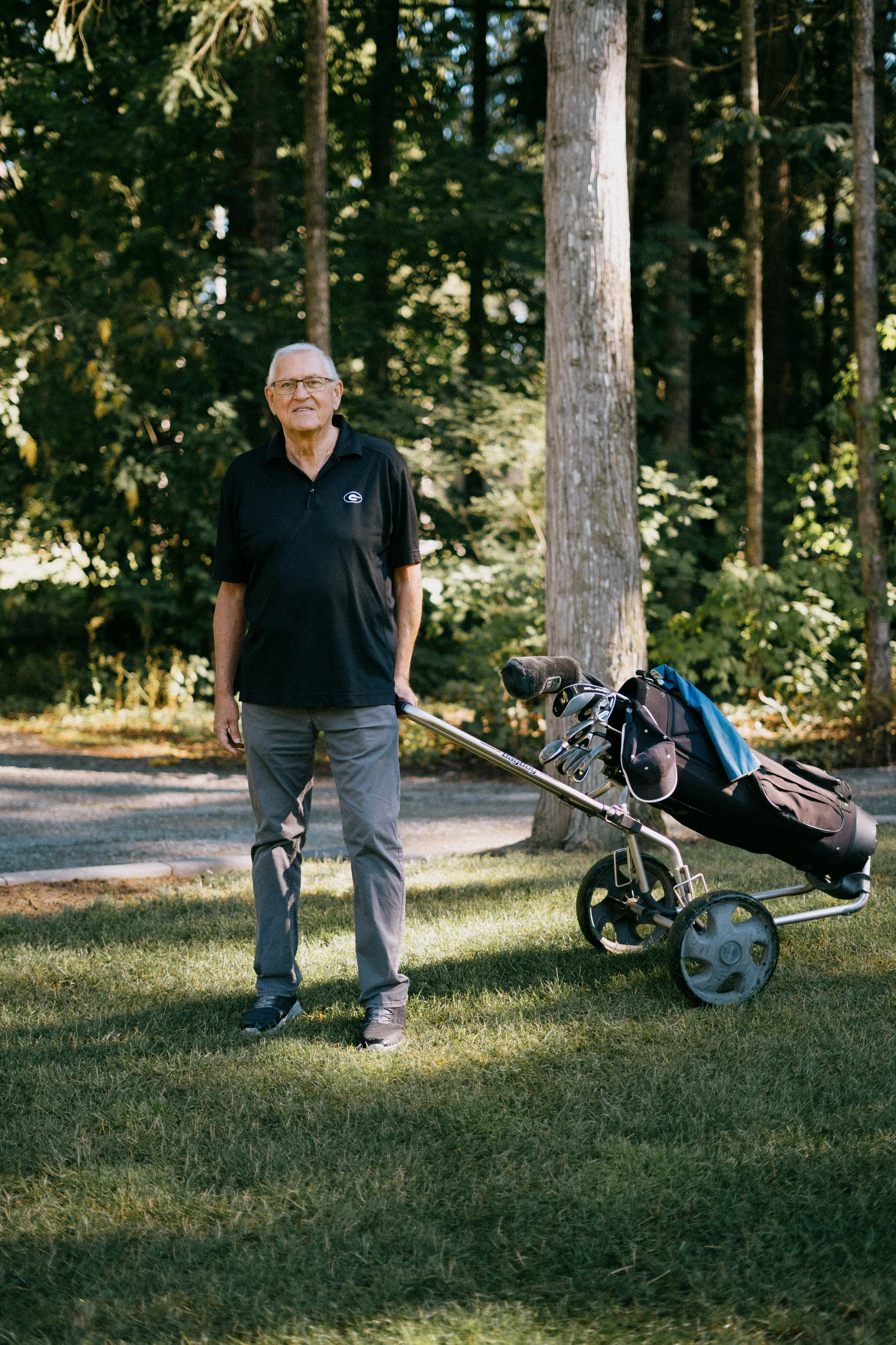 An elderly man in a black polo shirt and gray pants standing on a grassy area in a wooded park, holding the handle of a golf cart with golf clubs, with trees in the background. Photo by Vincent Prograce
