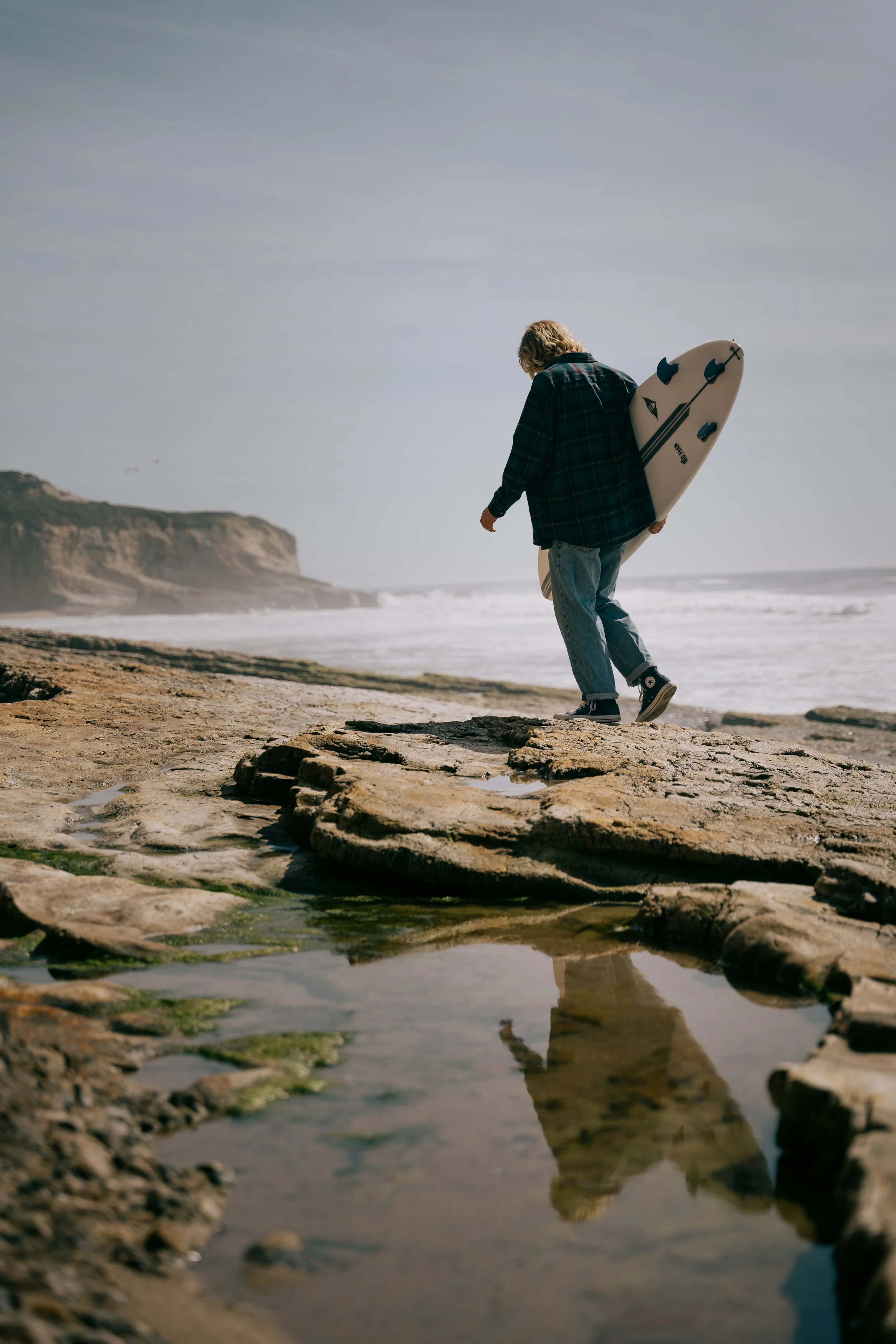A person walking along a rocky beach carrying a surfboard under their arm, with a distant shoreline and canyon in the background. Photo by Vincent Prograce