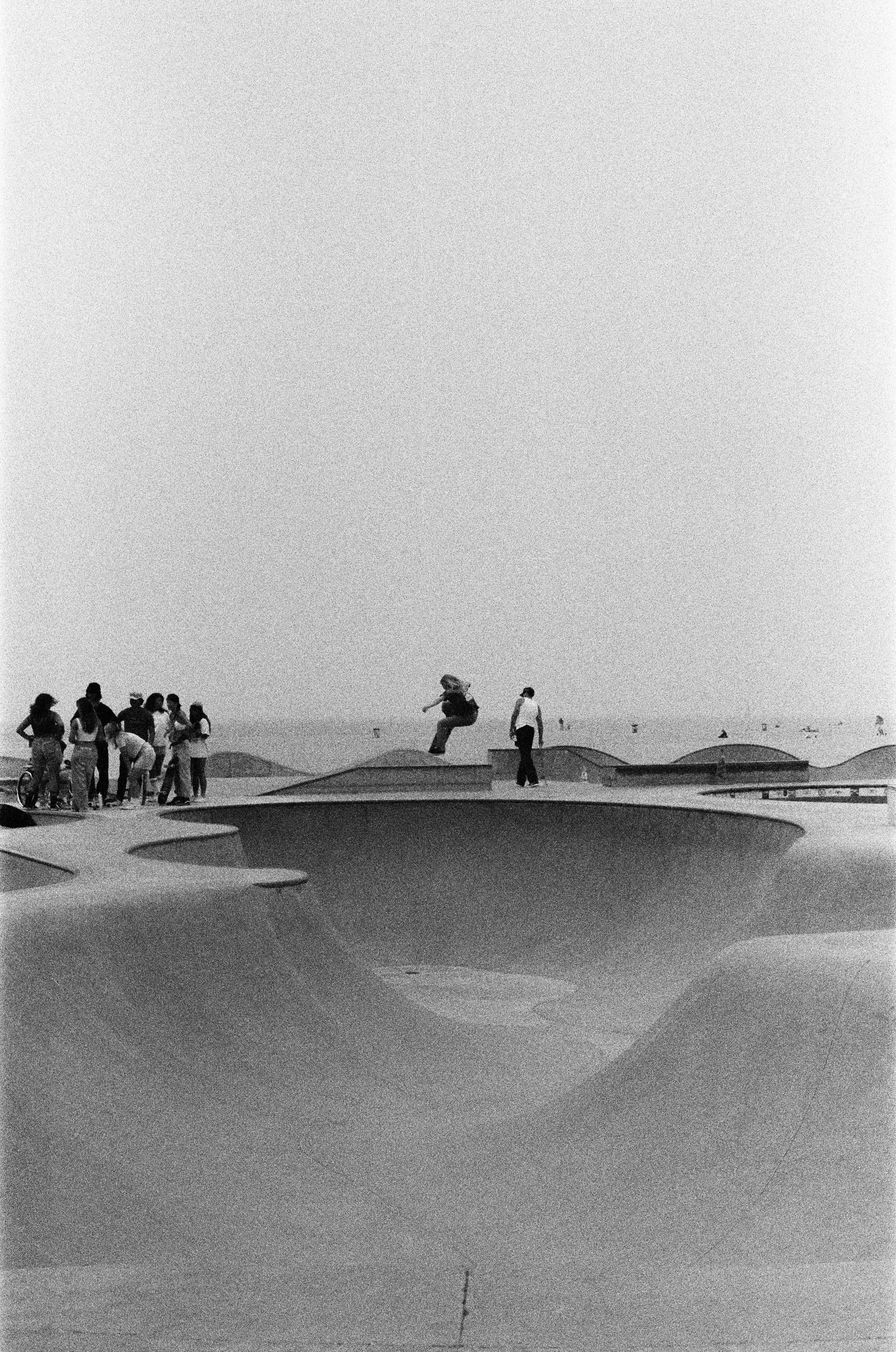 People at a skate park with concrete bowls, some standing and one person mid-air skateboarding. Photo by Vincent Prograce
