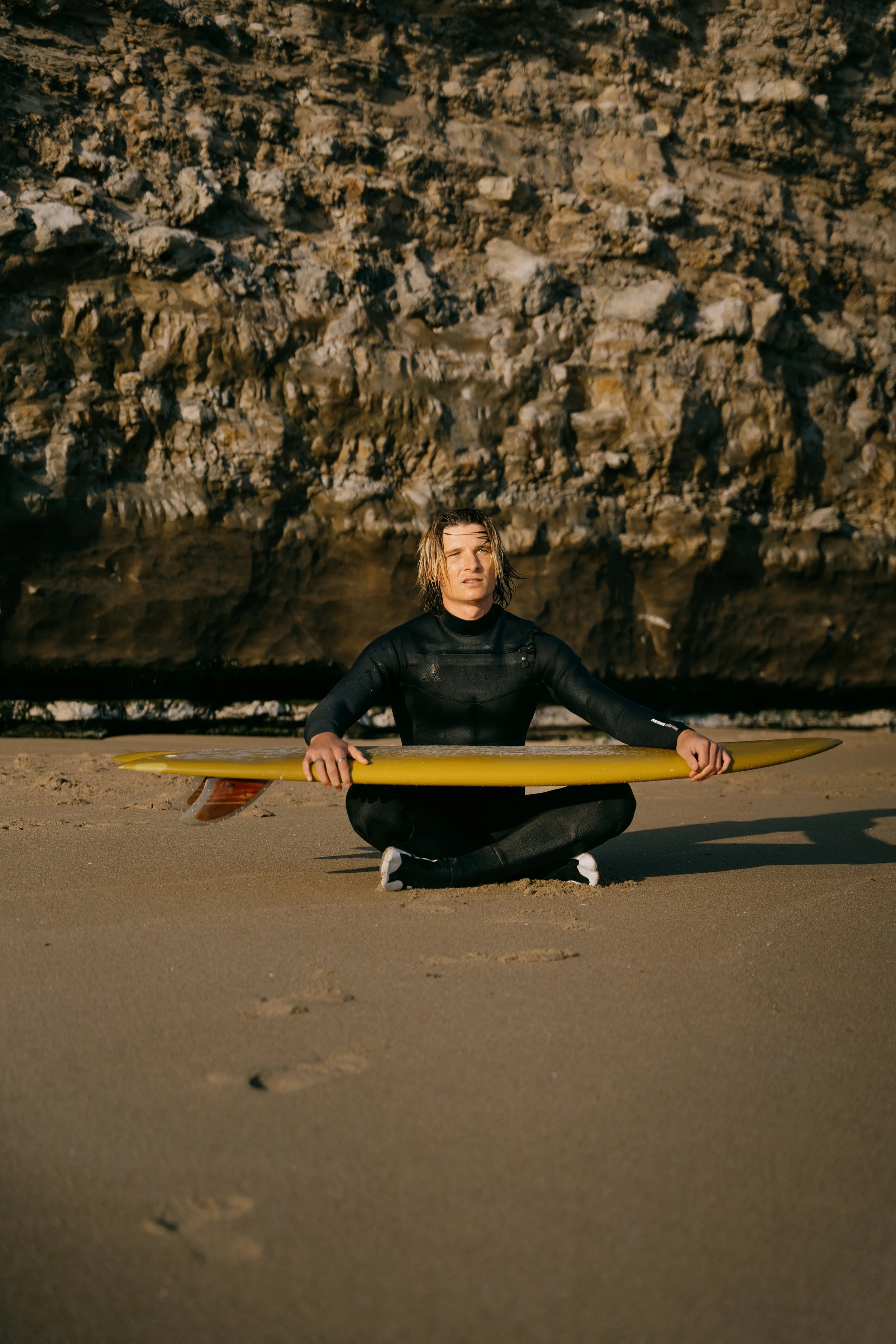 A male surfer in a wetsuit sitting cross-legged on sandy beach holding a yellow surfboard with fins. Photo by Vincent Prograce
