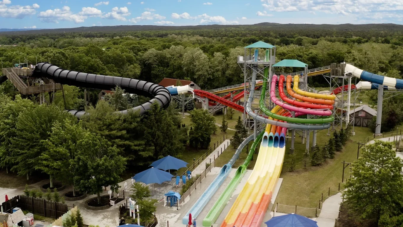 A water park with multiple colorful water slides and a large black enclosed slide, surrounded by green trees and a partly cloudy sky.