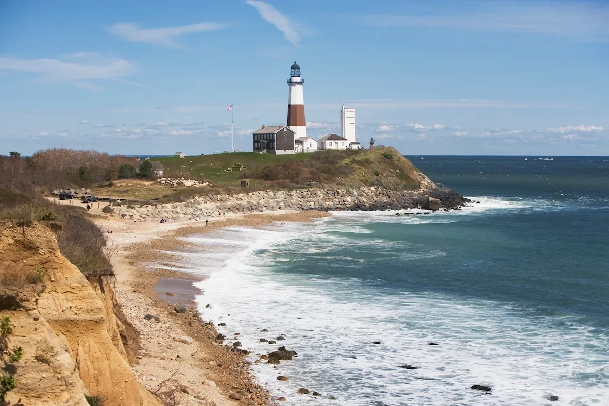 A coastal scene featuring a beach with rocky cliffs in the foreground, a lighthouse on a grassy hill, and the ocean with waves and a blue sky in the background.
