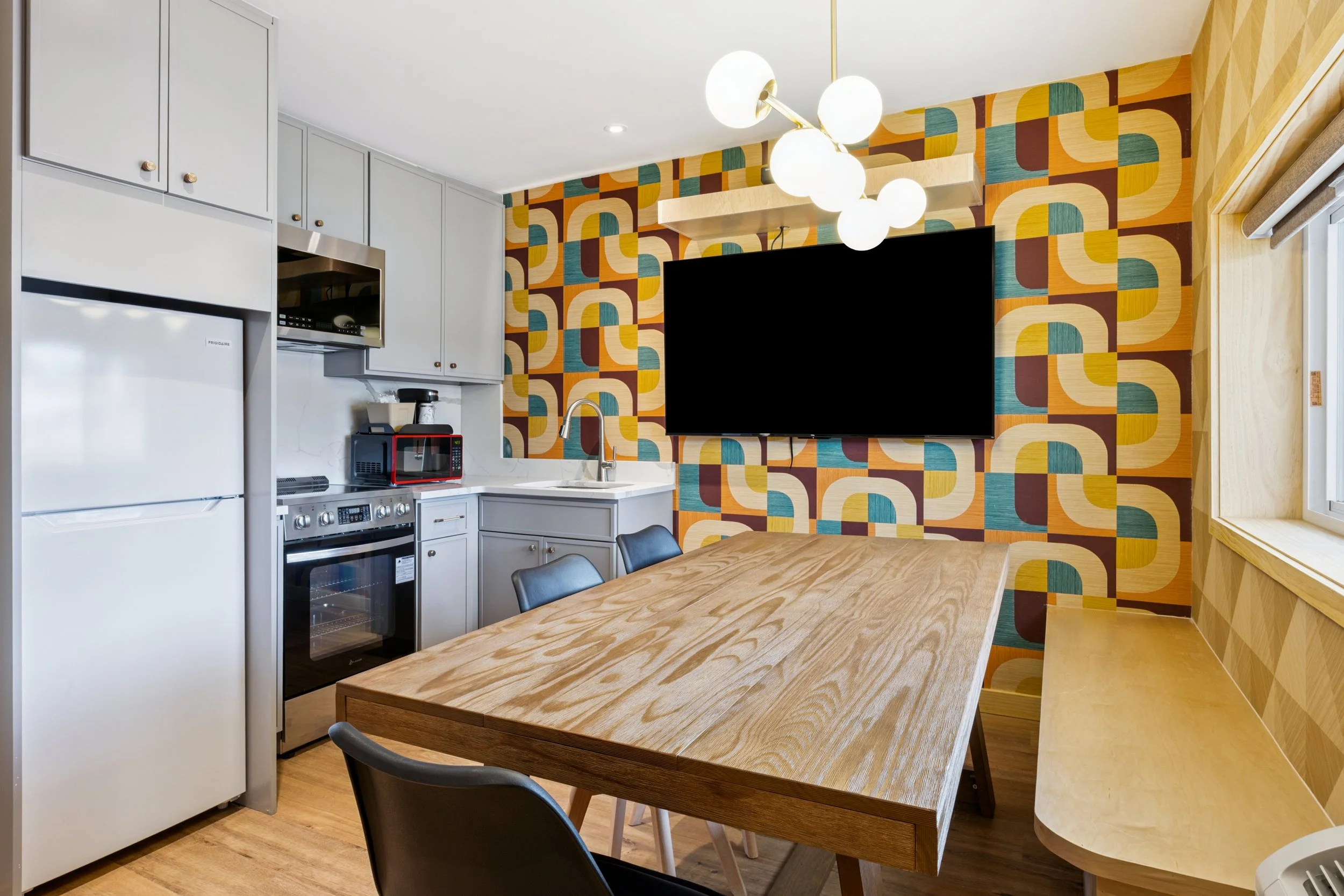 Kitchen and dining area with a wooden table, black chairs, colorful geometric wallpaper, a large black television, and kitchen appliances including a refrigerator, microwave, and oven.