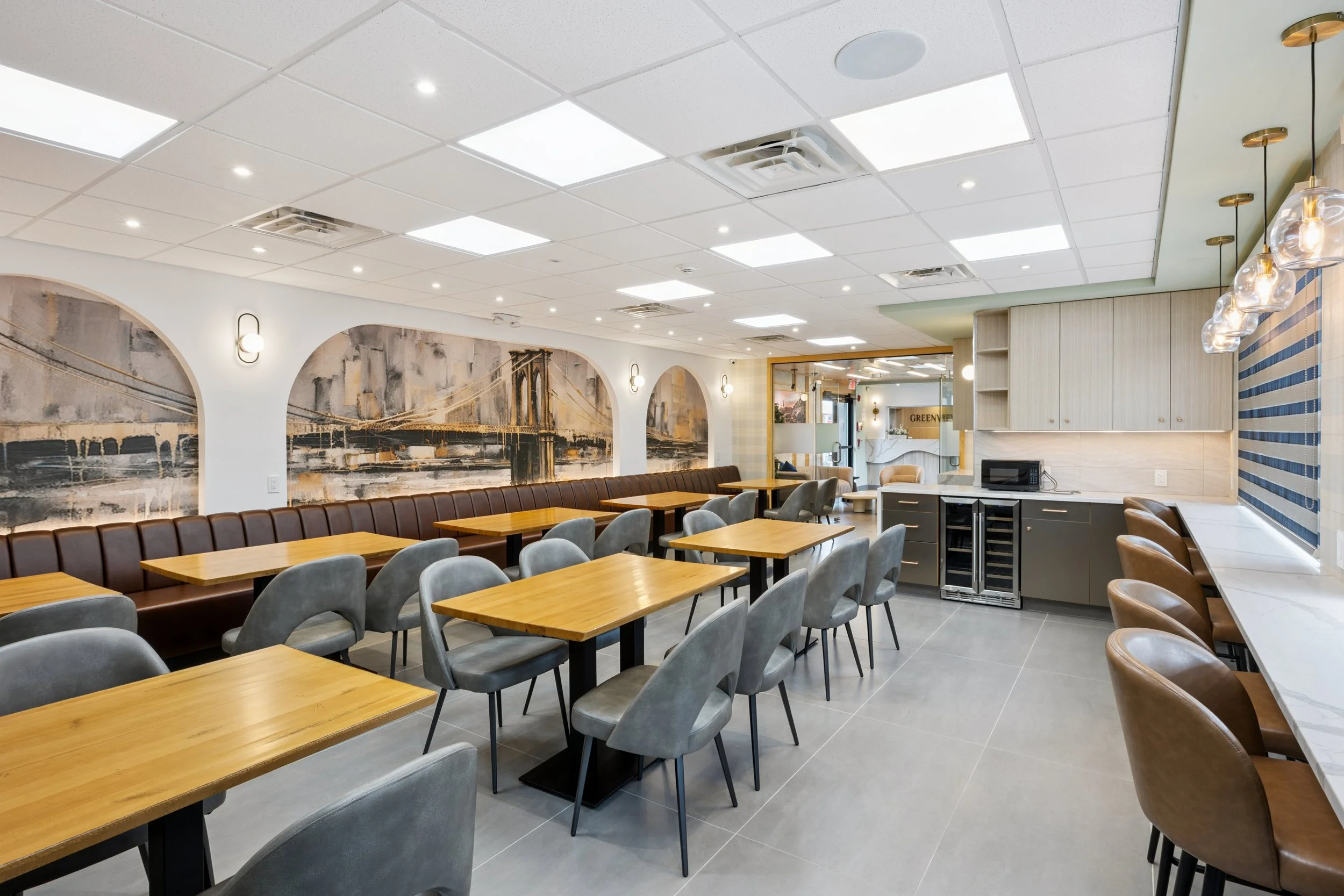 Modern indoor dining area with wooden tables, gray and brown chairs, wall art depicting bridges, and a counter with microwave and mini fridge.