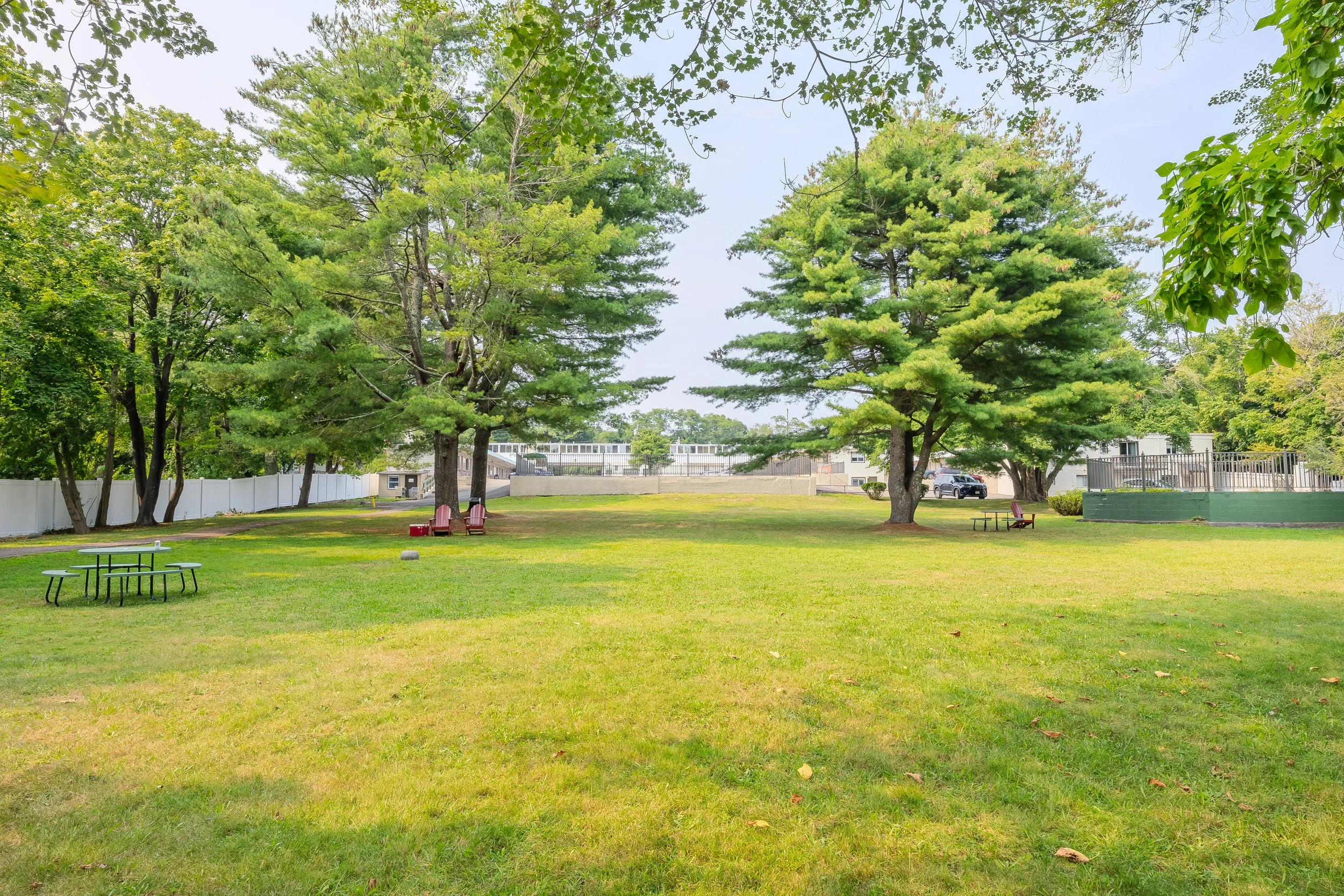 A grassy park with large green trees, picnic tables, and benches, enclosed by a white fence and some buildings in the background.