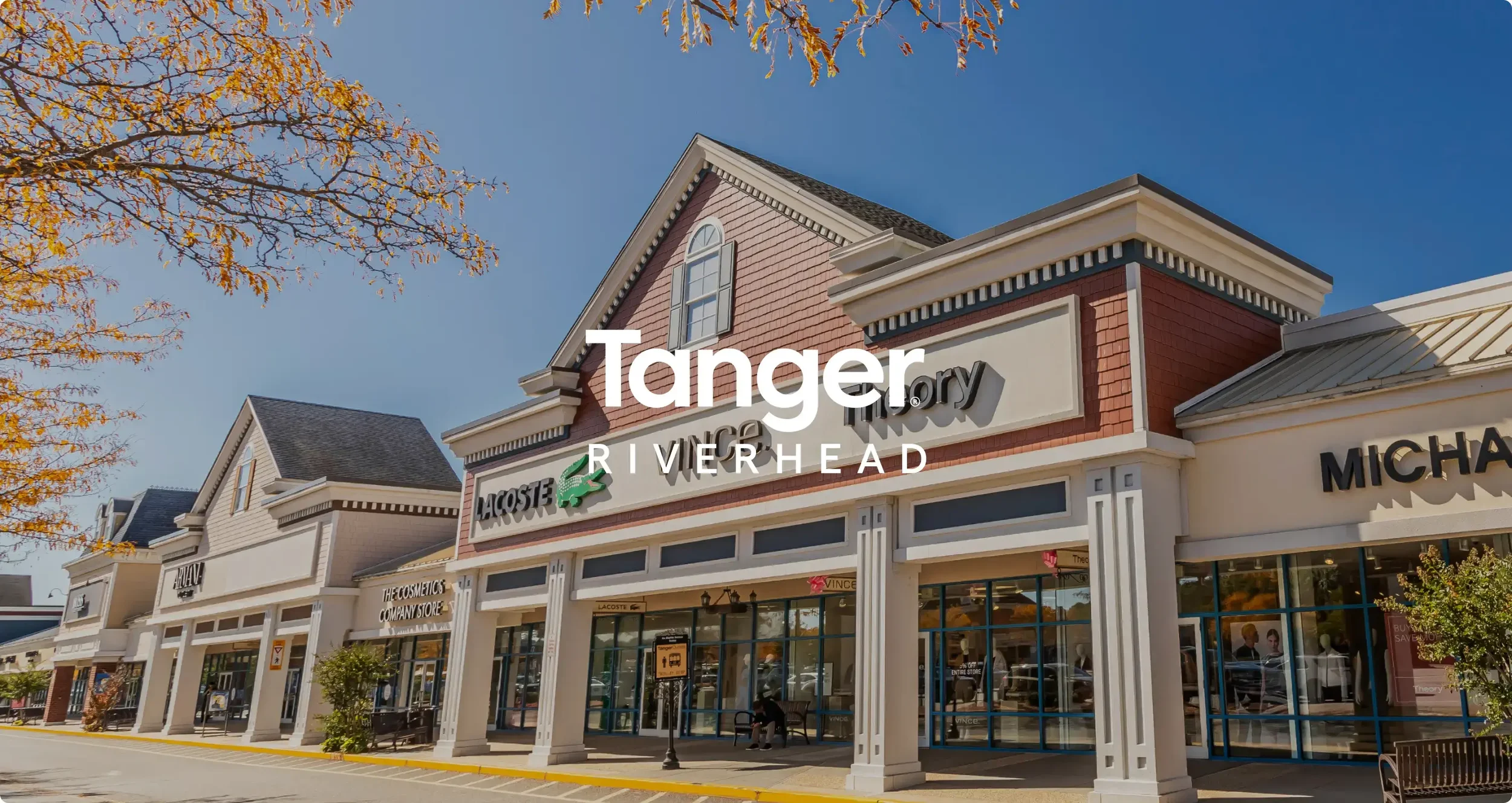 Retail shopping plaza in Tanger Riverhead with stores including Lacoste, Michael Kors, and The Cosmetics Company Store, under a clear blue sky with fall foliage.