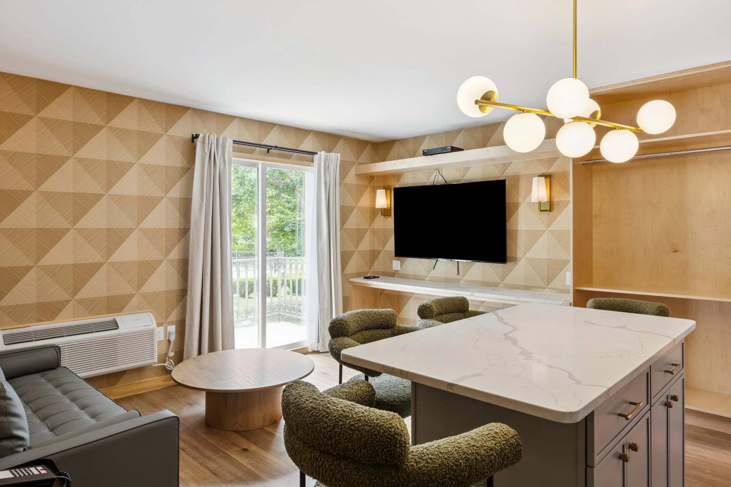 Living room with a large flat-screen TV, green upholstered chairs, a marble-topped kitchen island, a round coffee table, a sliding glass door with white curtains, and modern light fixtures.