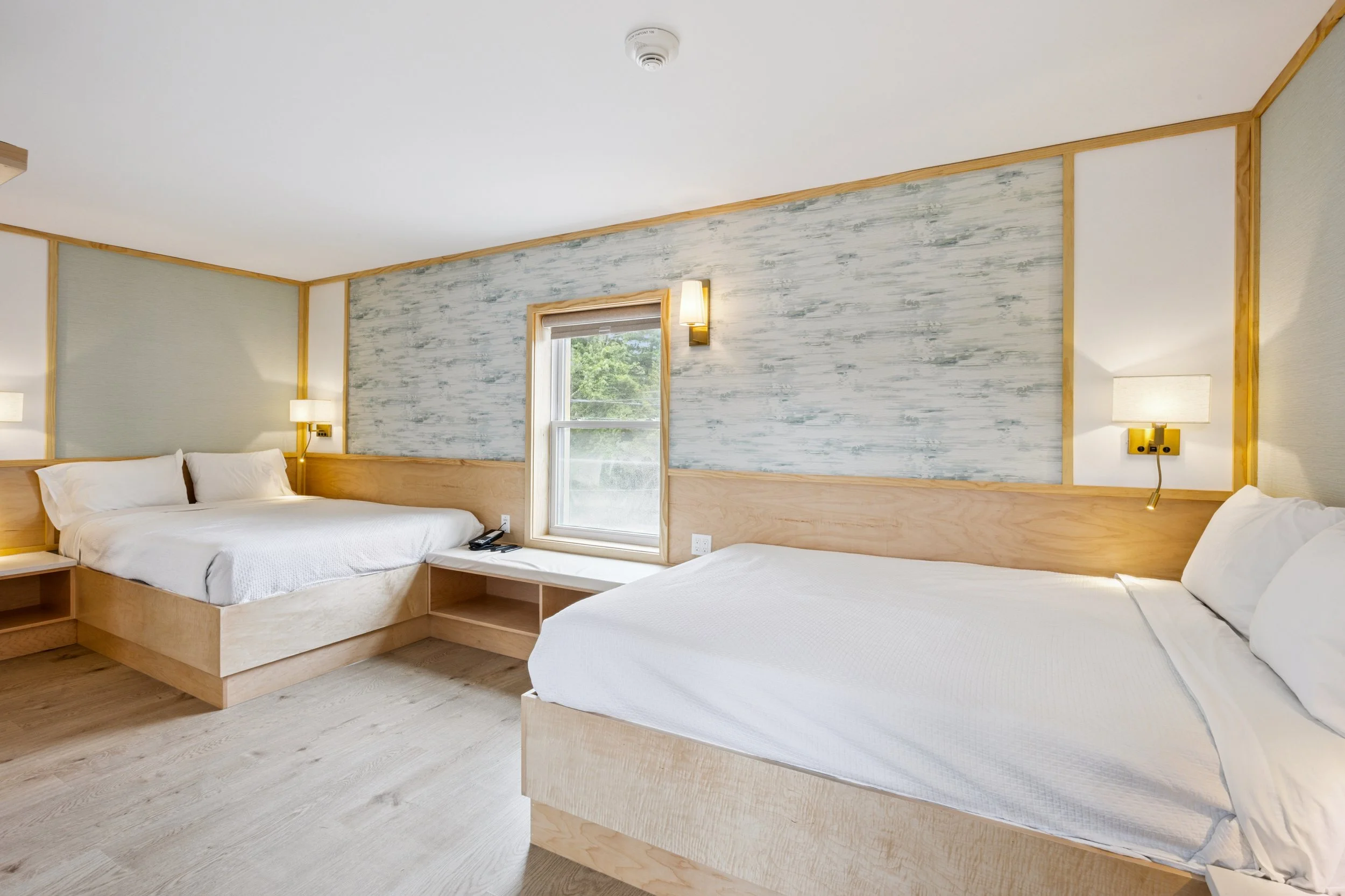 A hotel room with two beds, wood accents, a window with a view of greenery, and modern lighting fixtures.