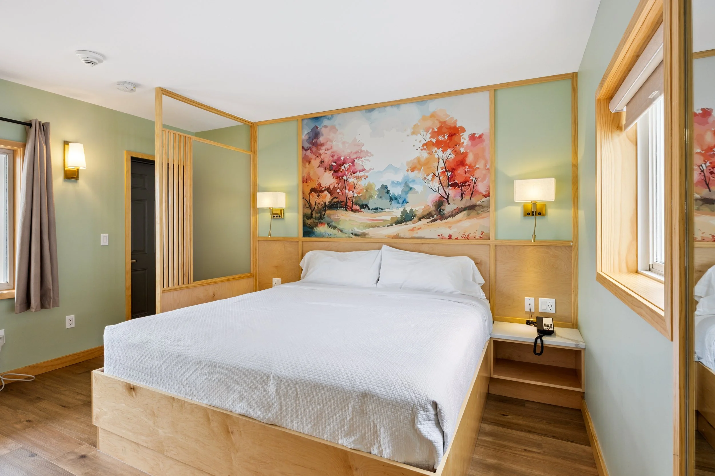 Hotel room with a bed, wooden accents, a colorful mountain landscape painting, and two wall-mounted lamps.