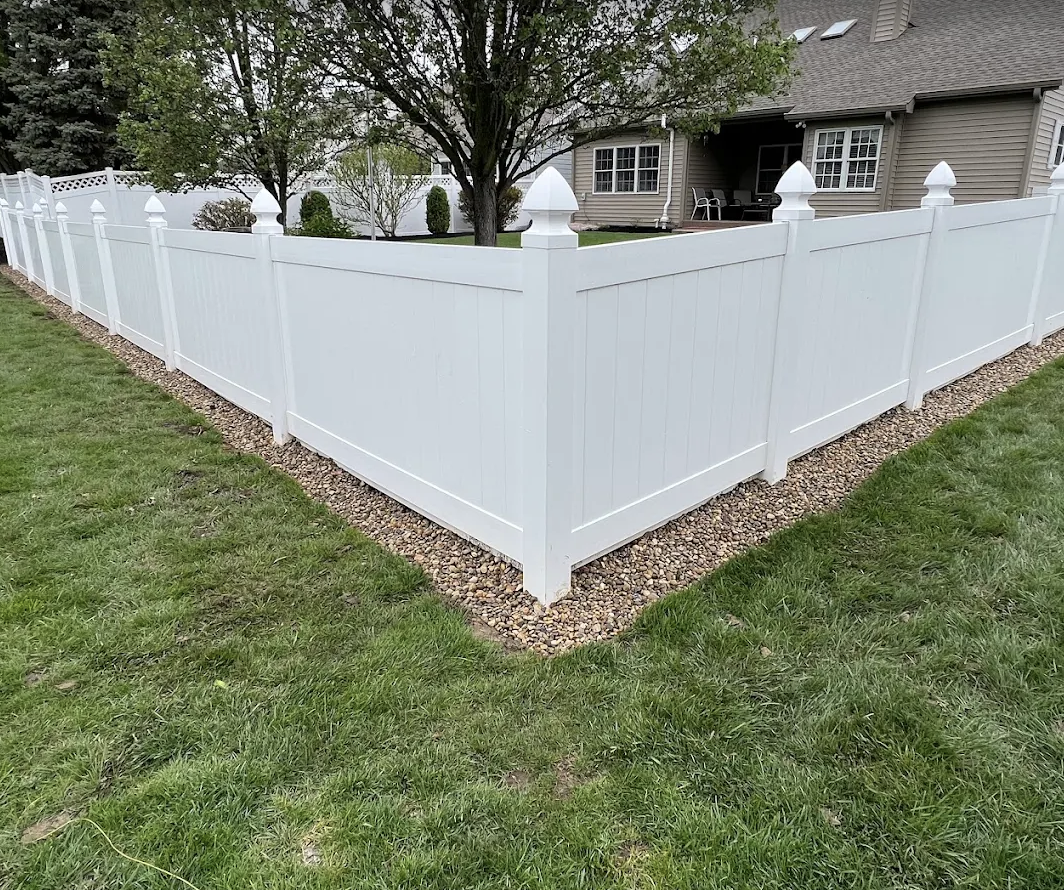A white vinyl privacy fence surrounds a backyard with a house, trees, and a patio with outdoor furniture.