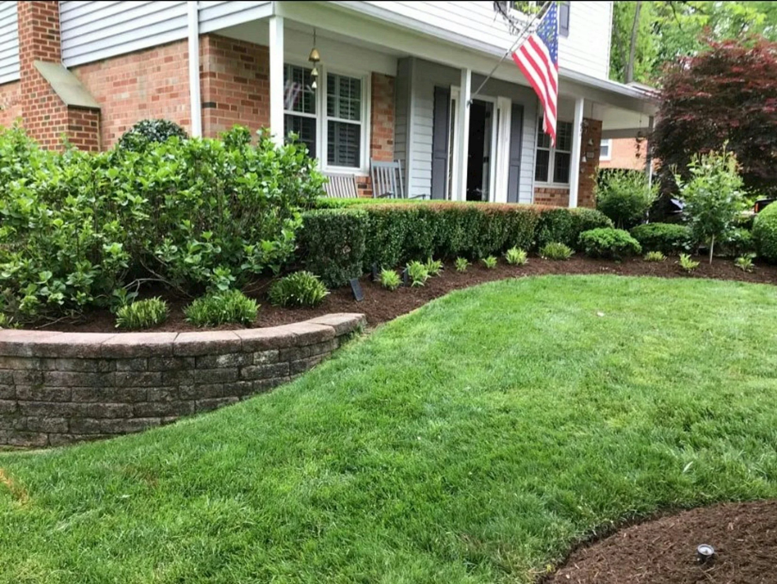 Front yard with well-maintained green grass, landscaped flower bed with various shrubs and plants, brick and siding house with porch, rocking chairs, and an American flag.