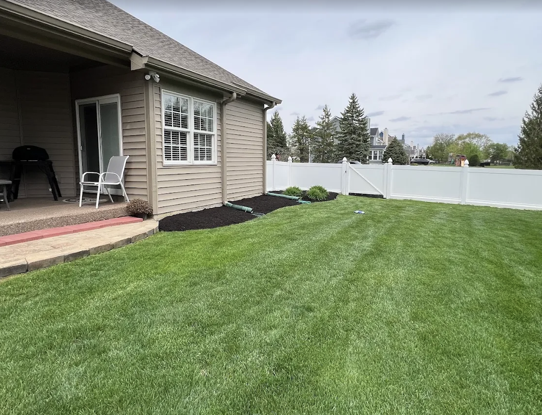 A backyard with a green lawn, a beige house with a porch, white chairs, and a white fence, with some landscaped garden beds and trees in the background.