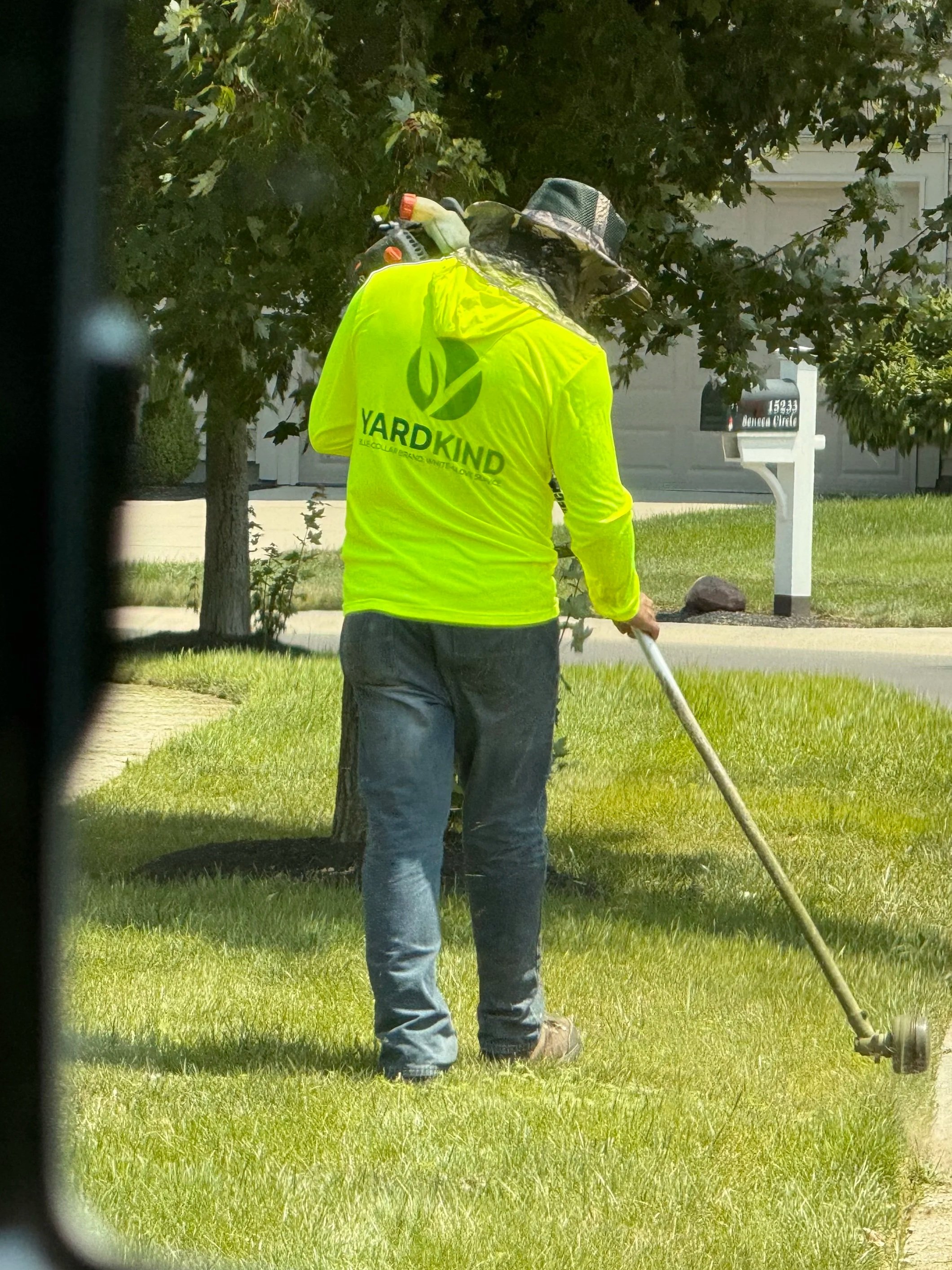 A man in an orange shirt is riding a riding lawn mower on a well-manicured green lawn in a residential backyard with trees, bushes, and houses in the background.
