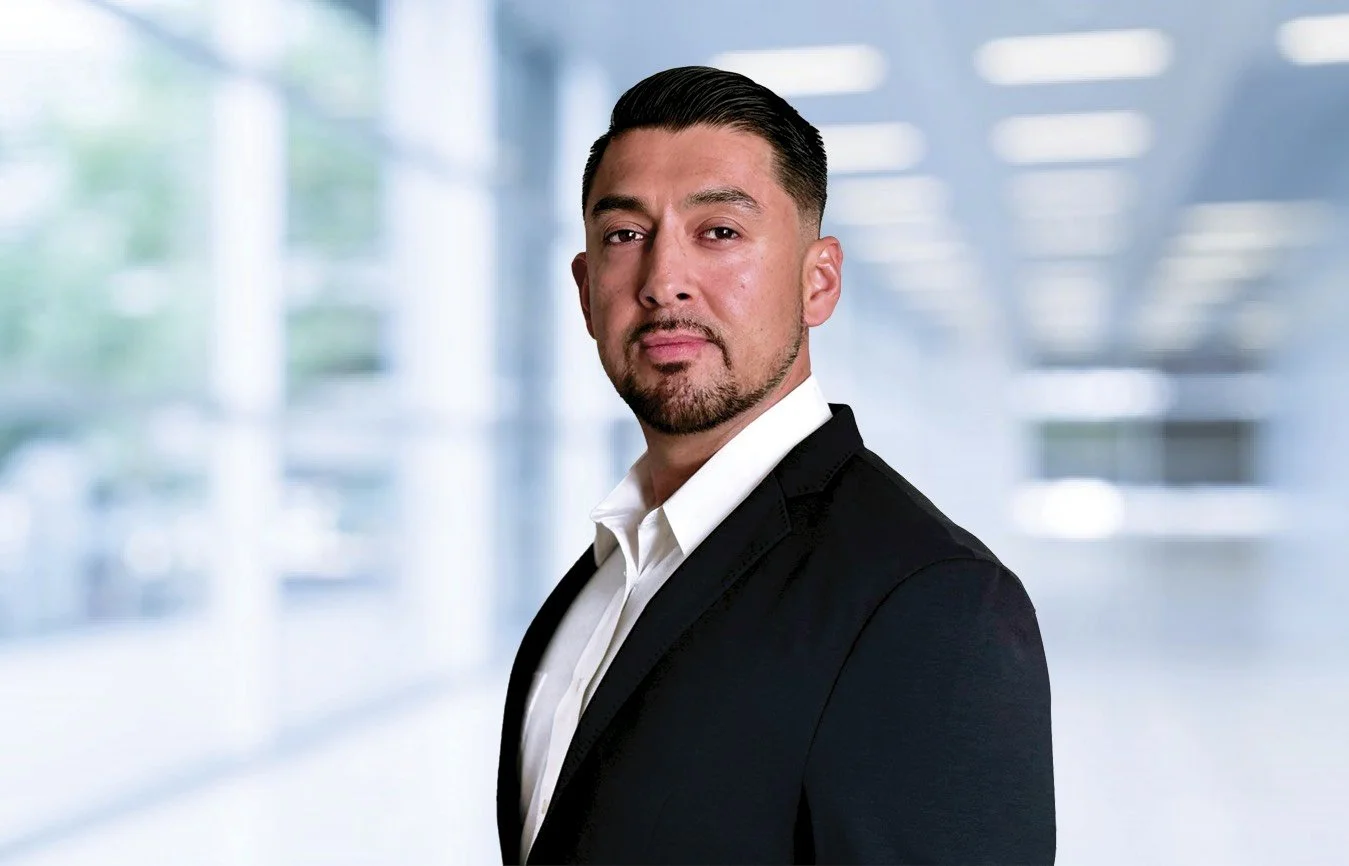 A professional man in a black suit with a white shirt in a modern office space with large windows and blurred background.