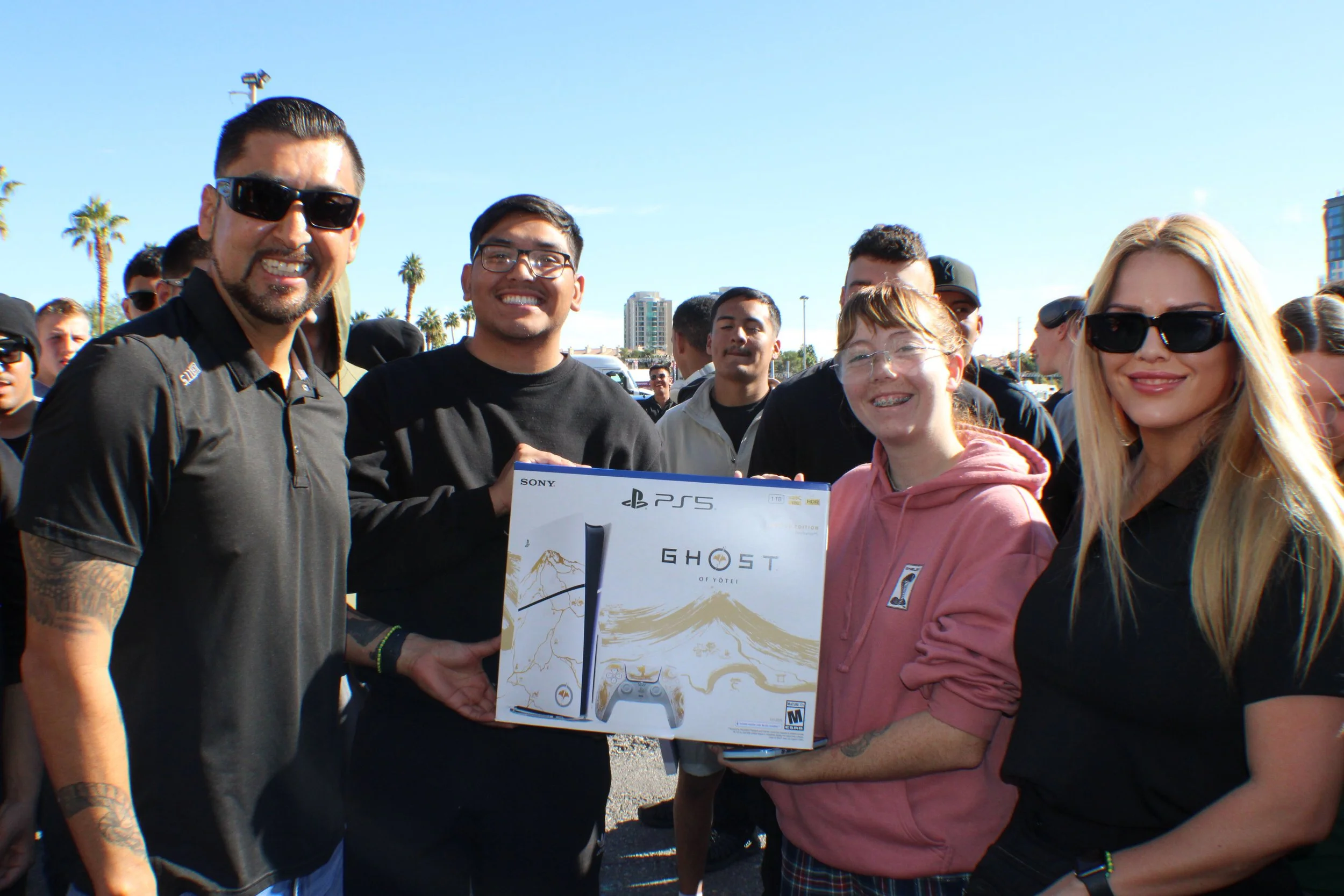 A person in military camouflage lifting a young girl in a navy dress with white long sleeves, laughing, at an outdoor patriotic event with American flags.